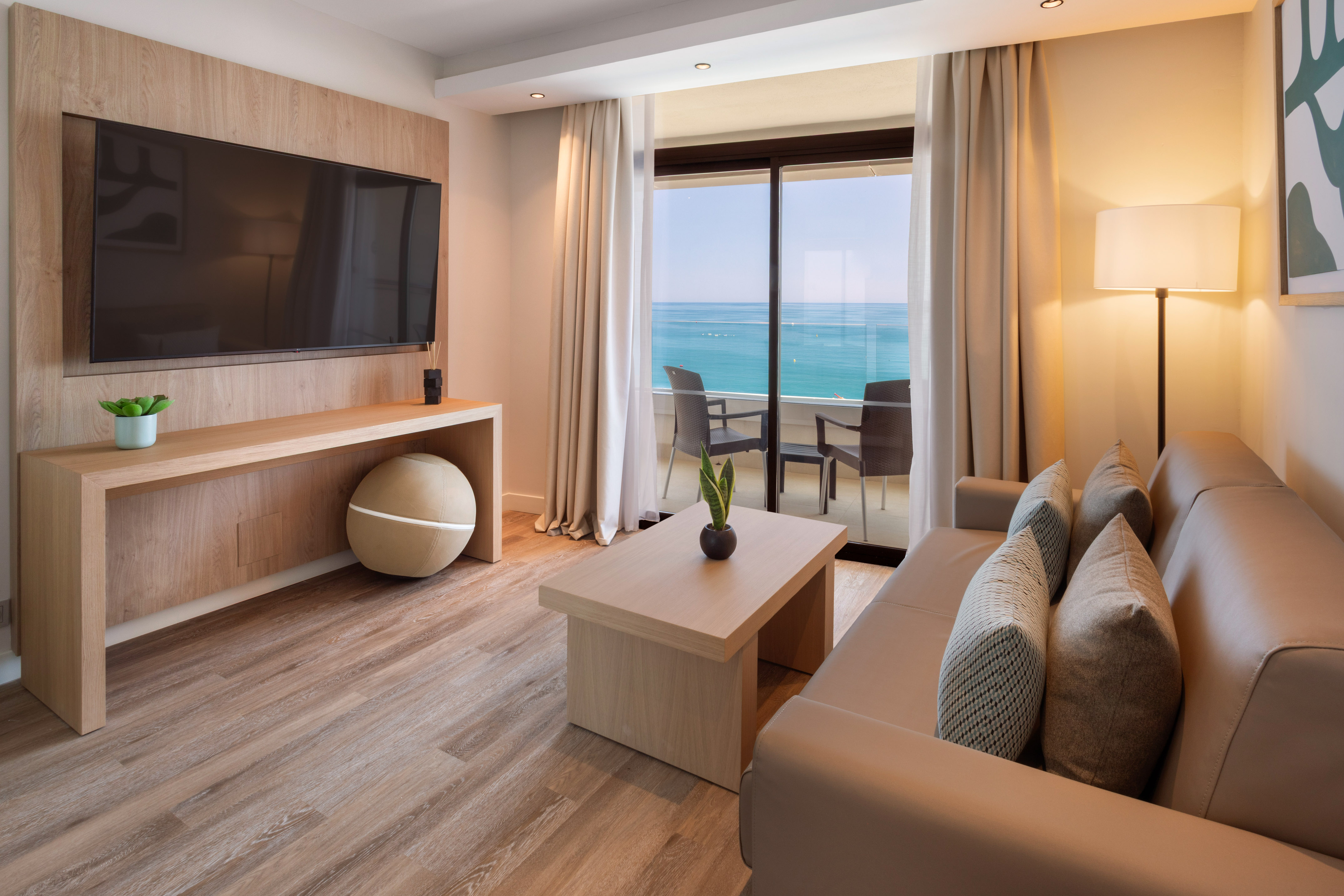 Hotel suite living room with ocean view balcony, modern sofa, and large TV.