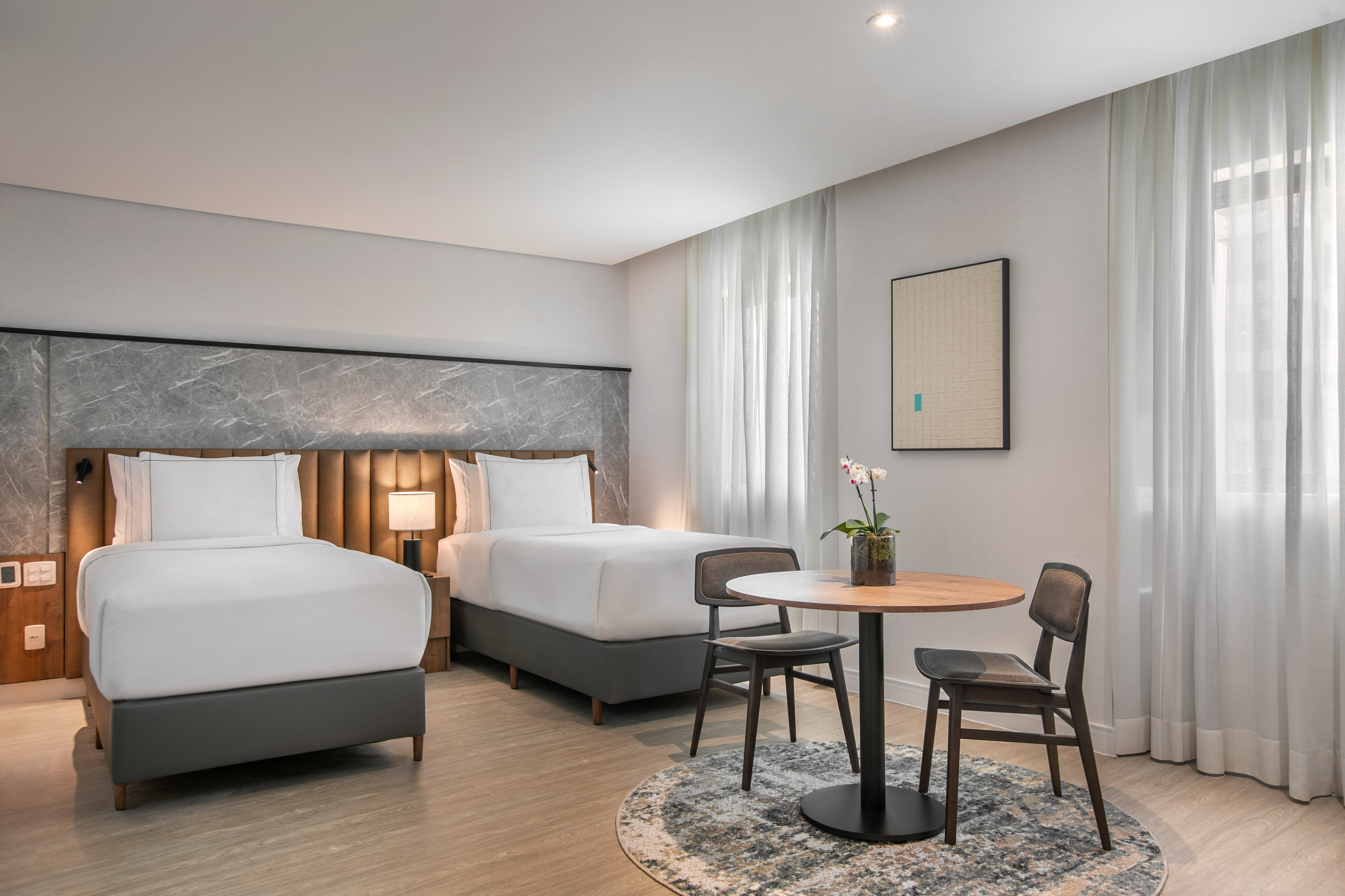 Modern hotel room with two twin beds, wood table, chairs, and marble headboard.