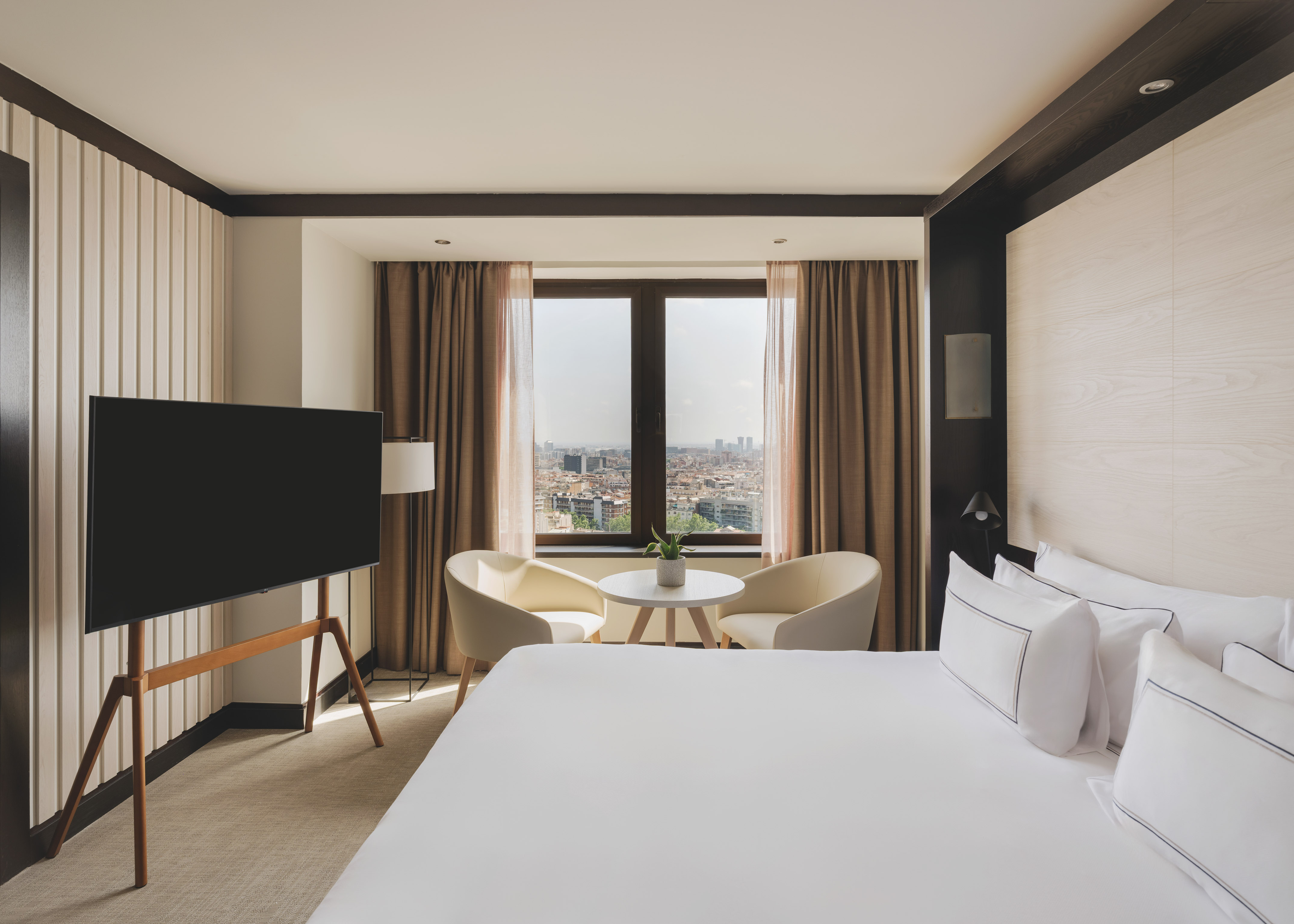 Habitación de hotel moderna con cama, TV, área de estar y vista urbana.