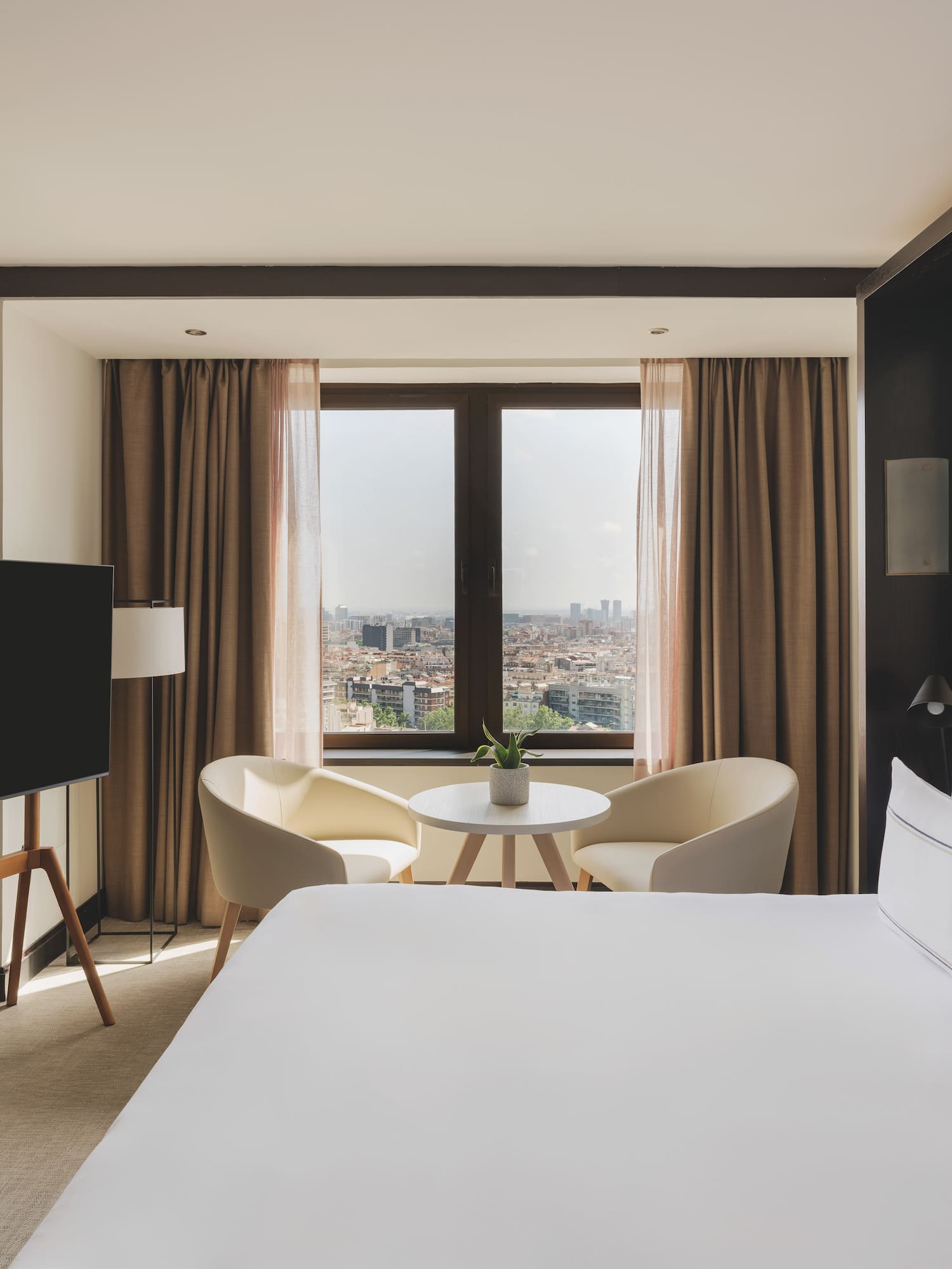 Modern hotel bedroom with city view, white bed, TV, and armchairs.