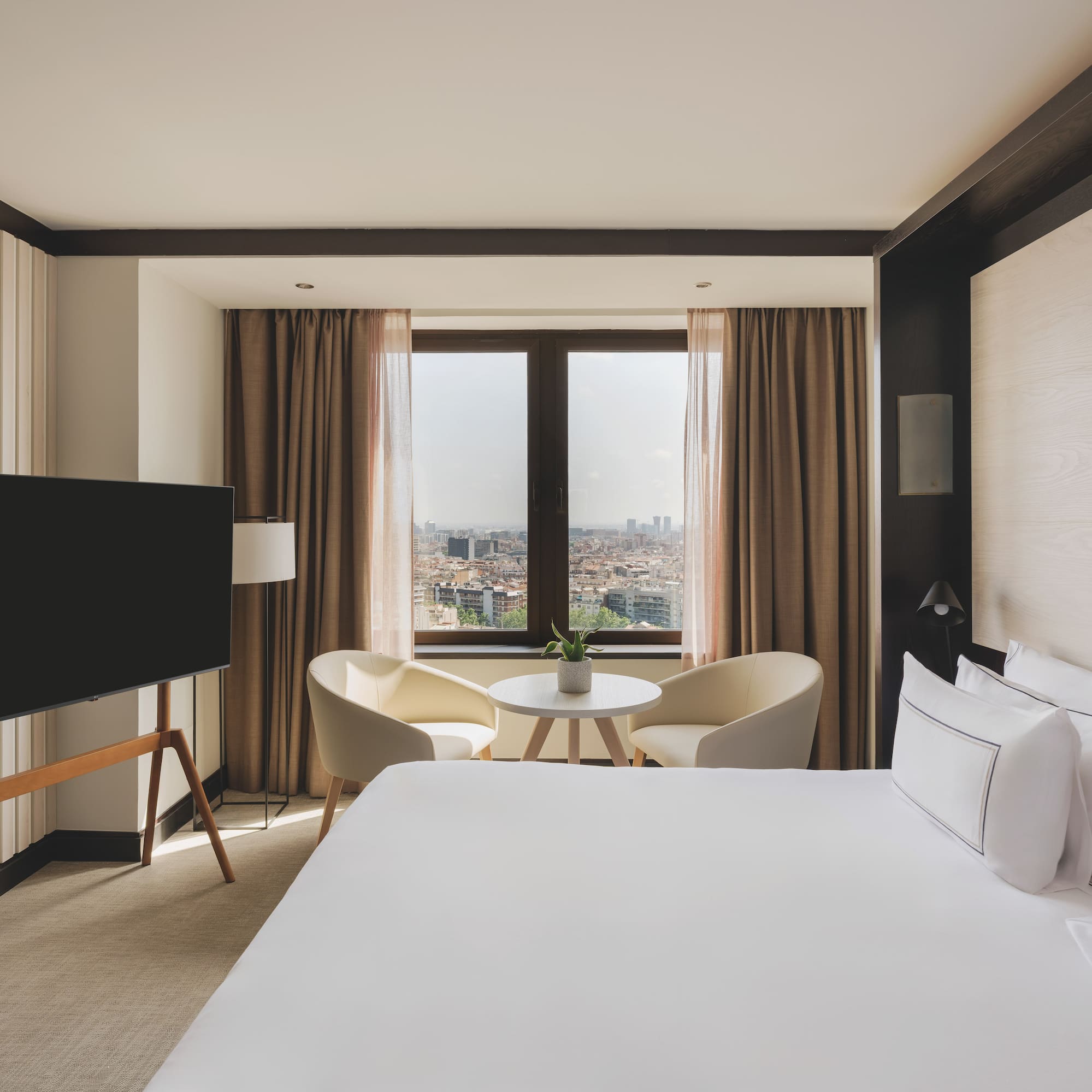 Habitación de hotel moderna con cama, TV, área de estar y vista urbana.