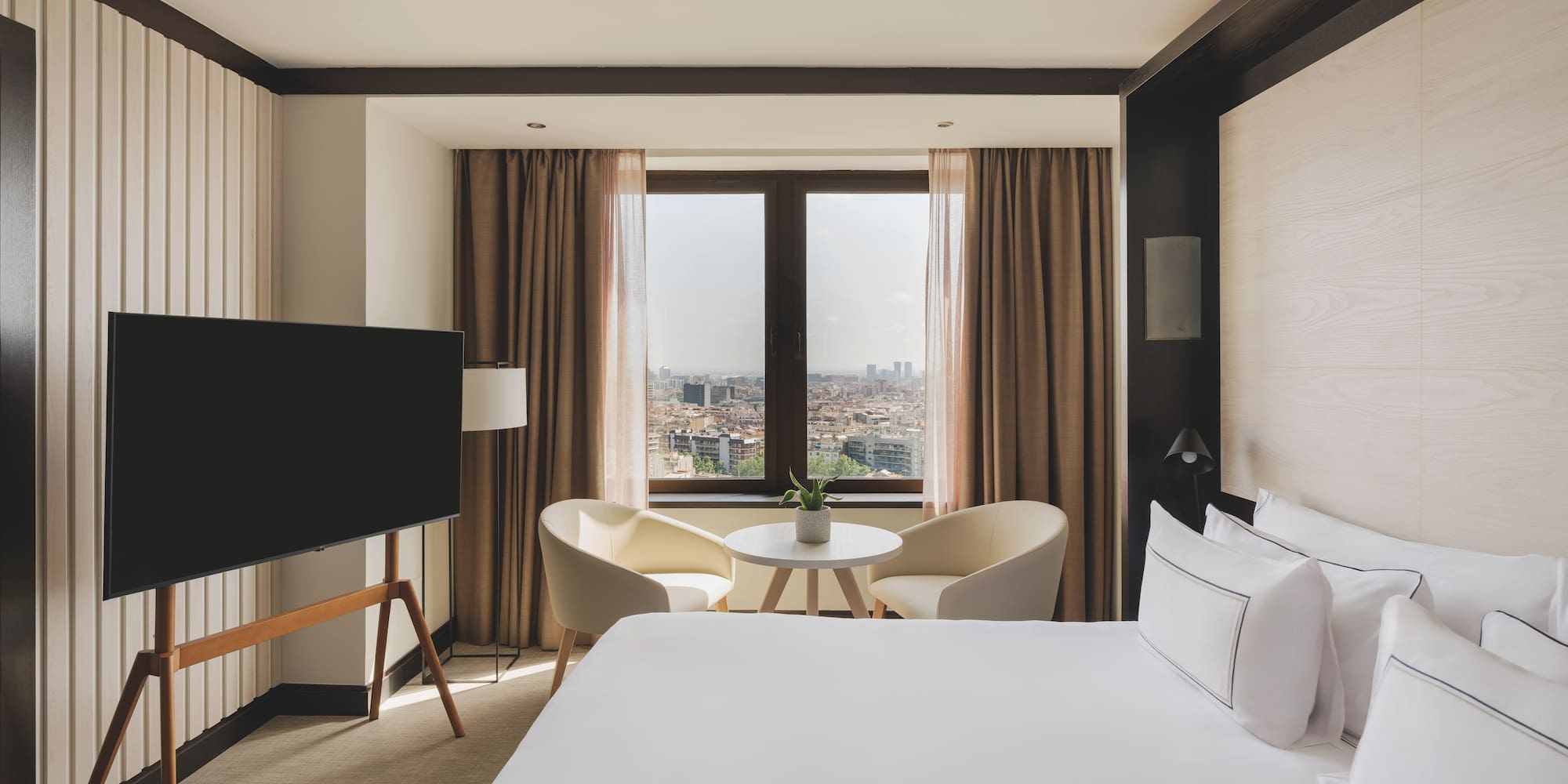 Habitación de hotel moderna con cama, TV, área de estar y vista urbana.