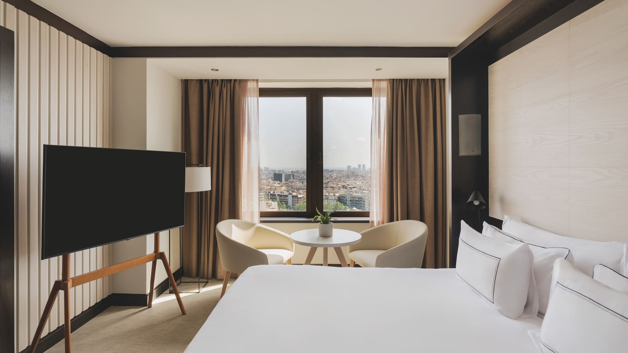 Habitación de hotel moderna con cama, TV, área de estar y vista urbana.