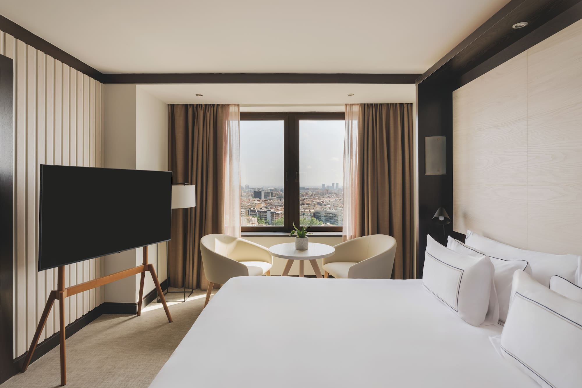 Habitación de hotel moderna con cama, TV, área de estar y vista urbana.