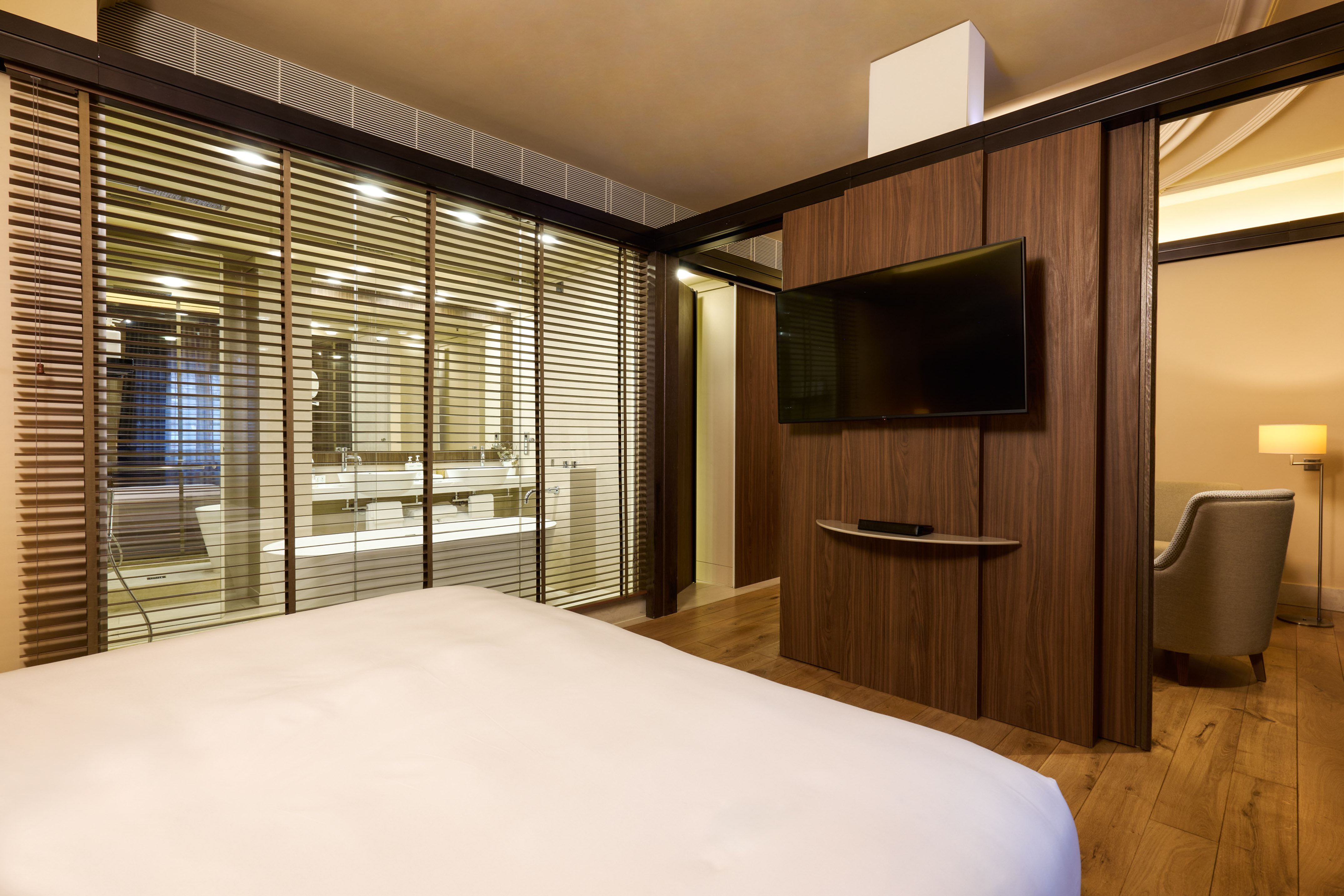 Modern hotel bedroom with glass bathroom, wooden blinds, and wall TV.