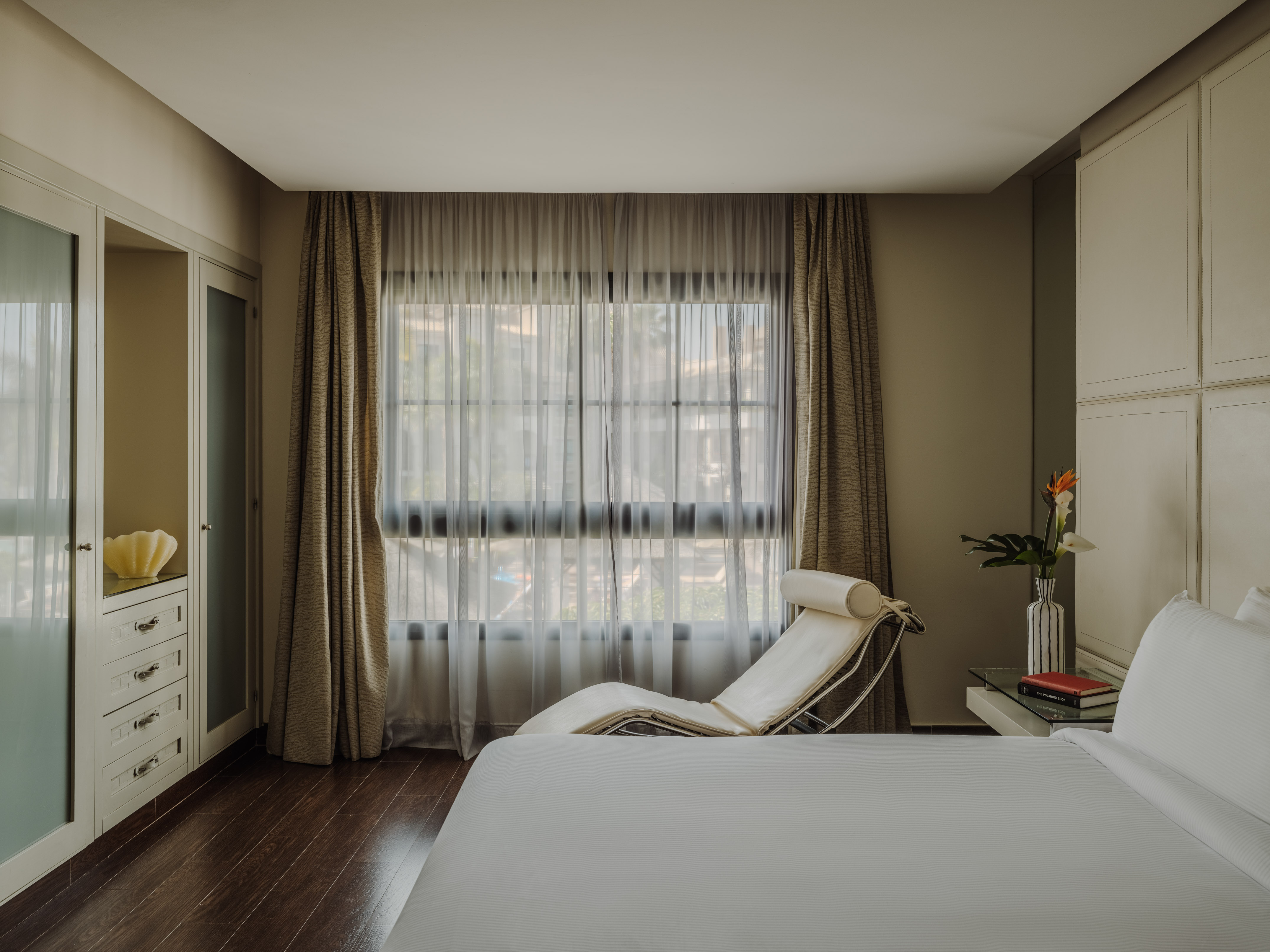 Modern bedroom with large window, white bed, chaise lounge, and storage.
