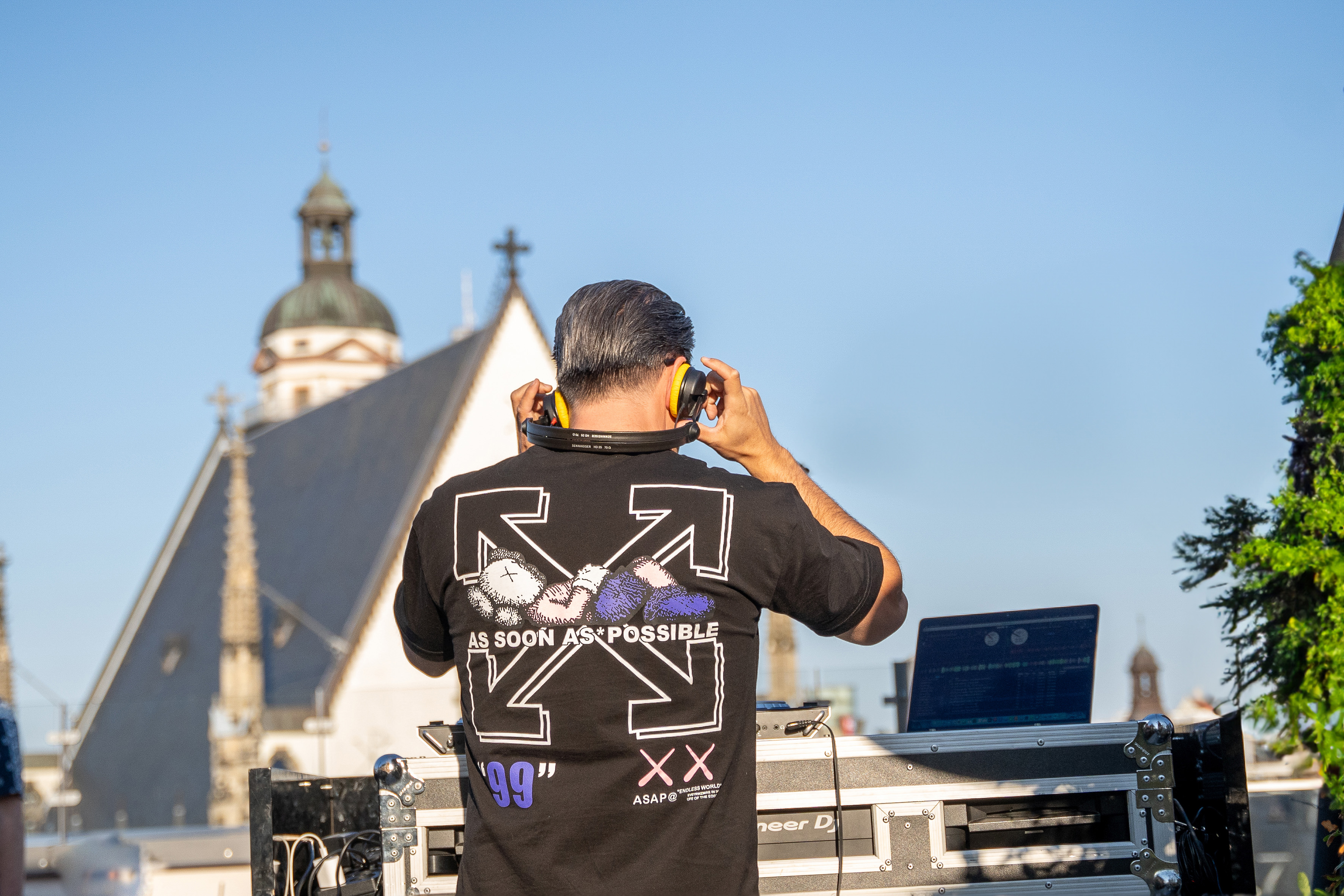 Rooftop DJ with headphones and graphic tee, overlooking a historic church.