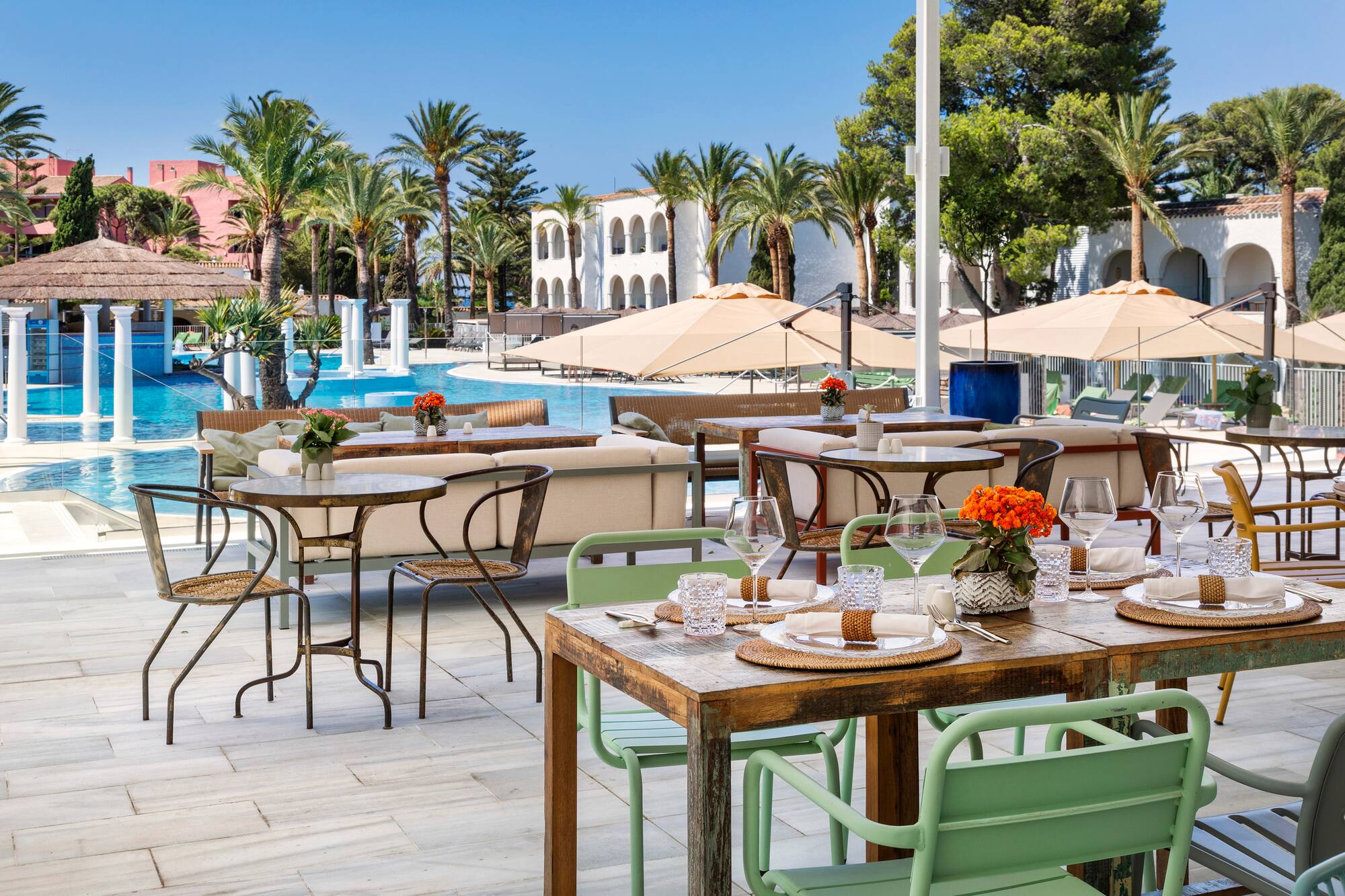 Sunny poolside restaurant with set dining tables, palm trees, and Mediterranean architecture.