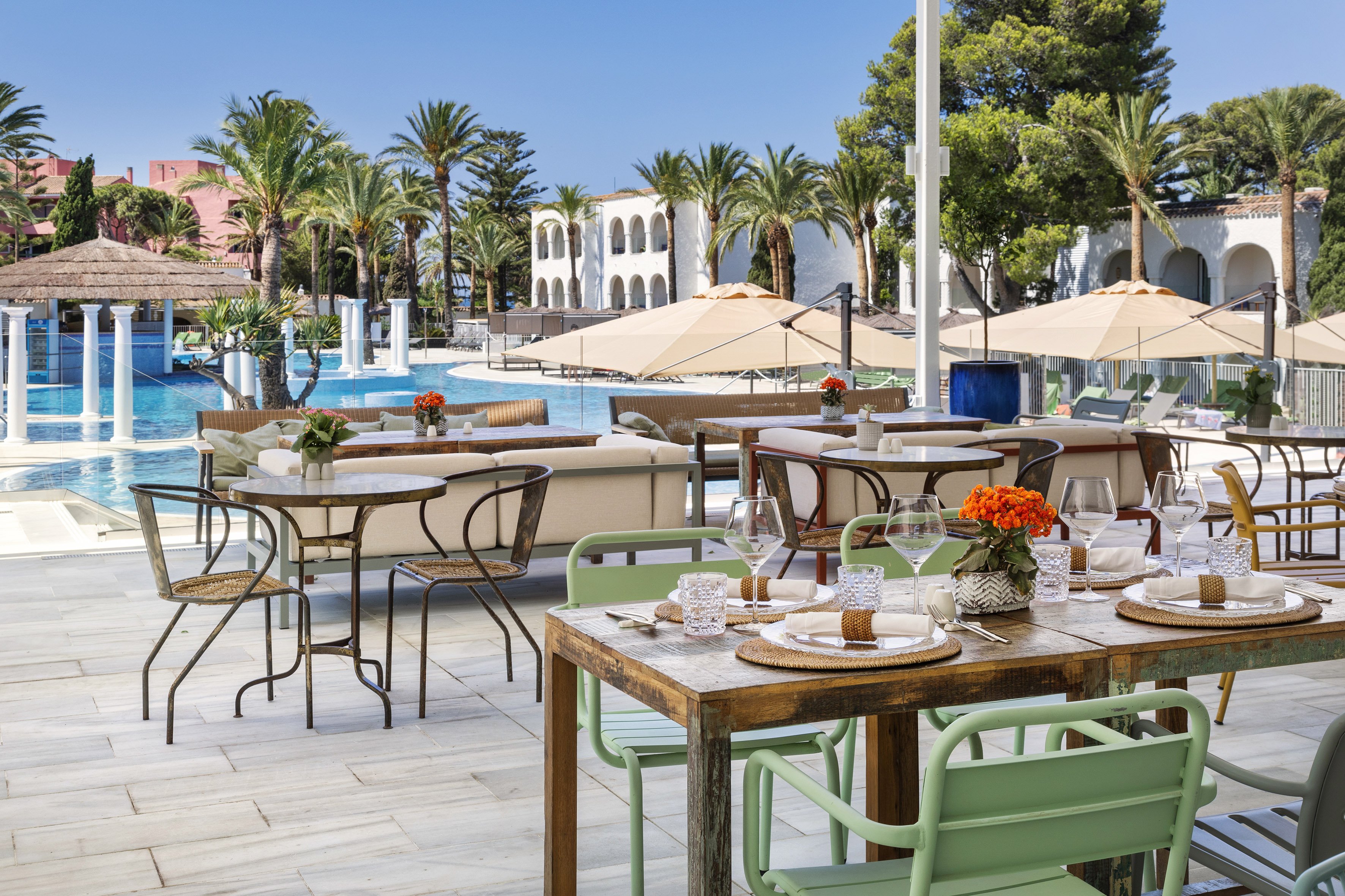 Sunny poolside restaurant with set dining tables, palm trees, and Mediterranean architecture.