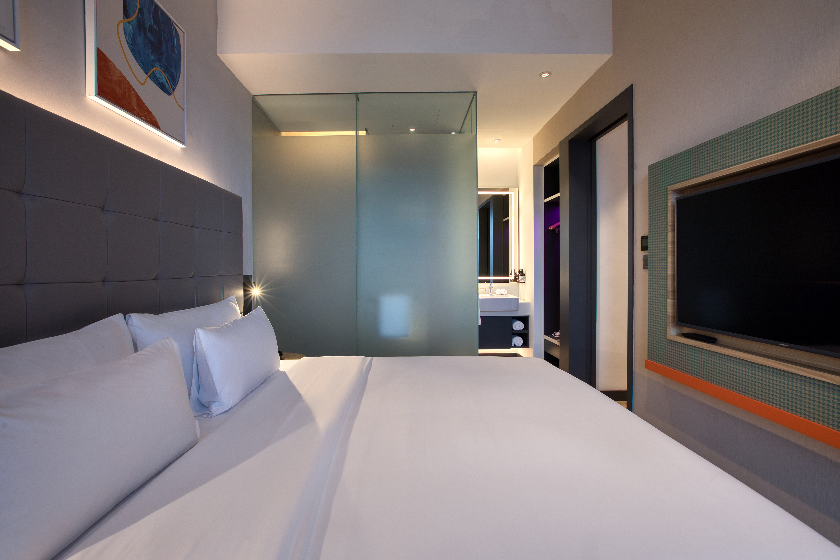 Modern hotel bedroom with king bed, frosted glass bathroom wall, and TV.