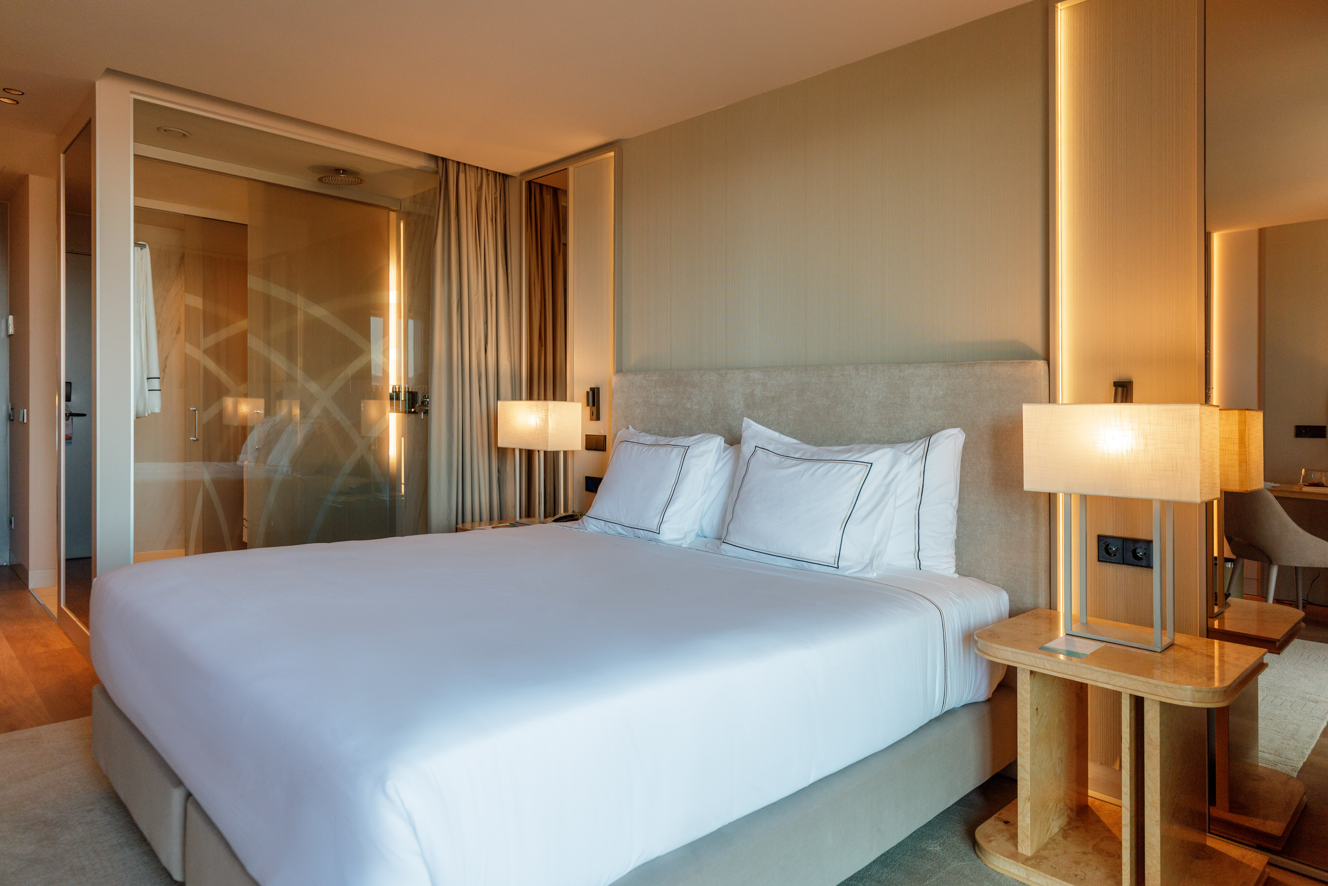 Modern hotel bedroom with white bed, glass bathroom, and elegant decor.