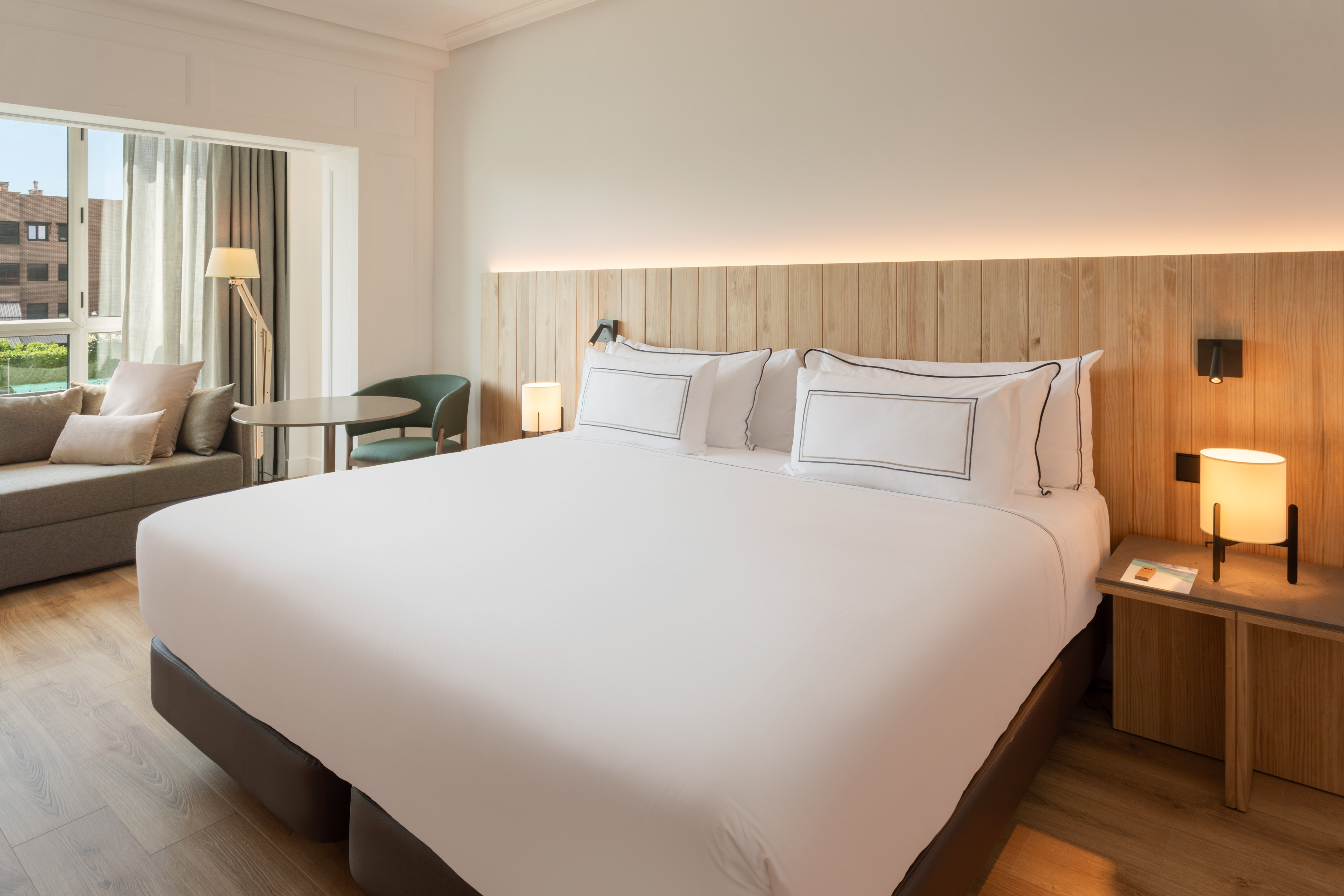 Modern hotel bedroom with large bed, wooden headboard, sofa, and window view.