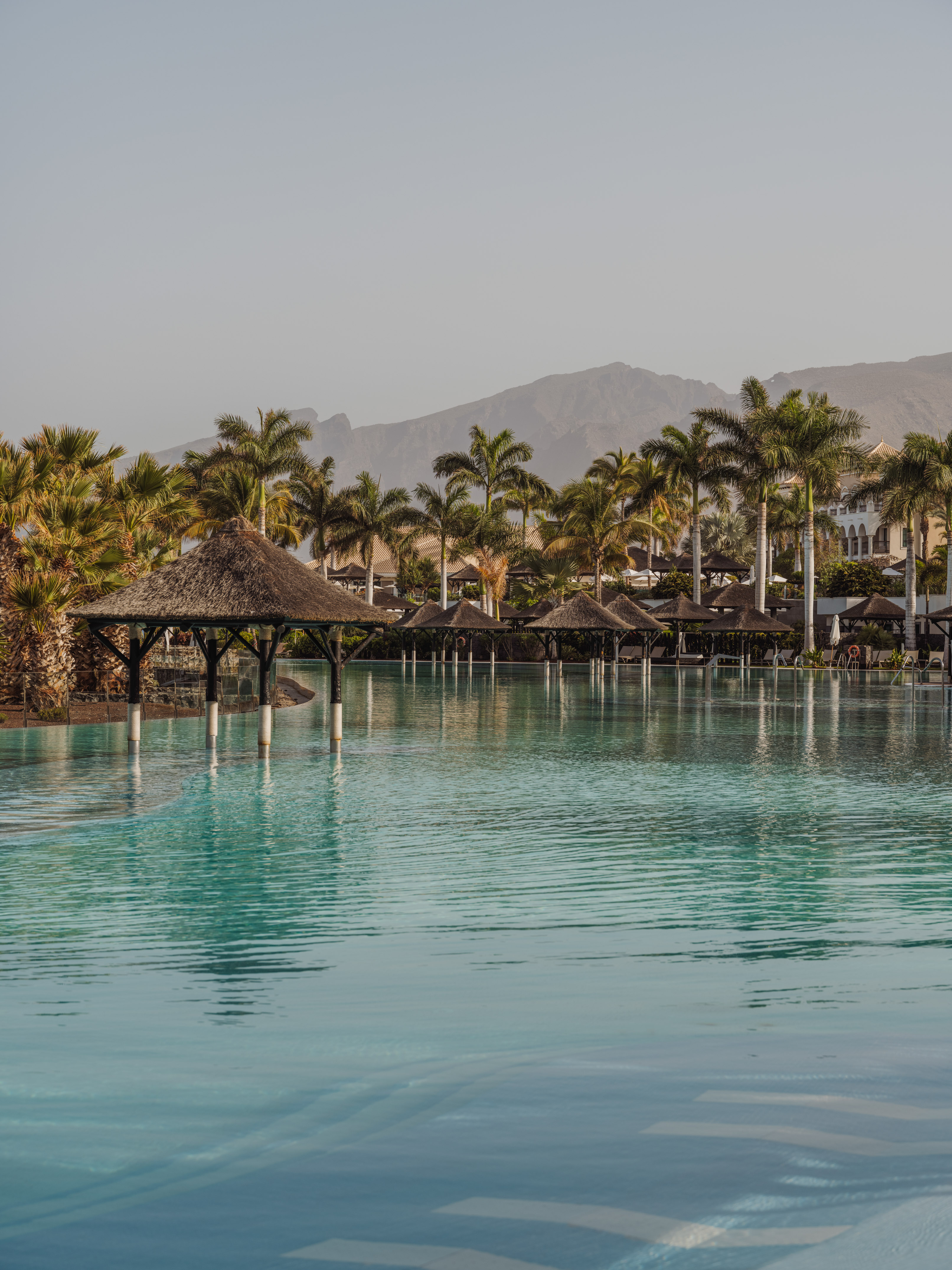 Tropical resort pool with palm trees, thatched palapas, and mountain views.
