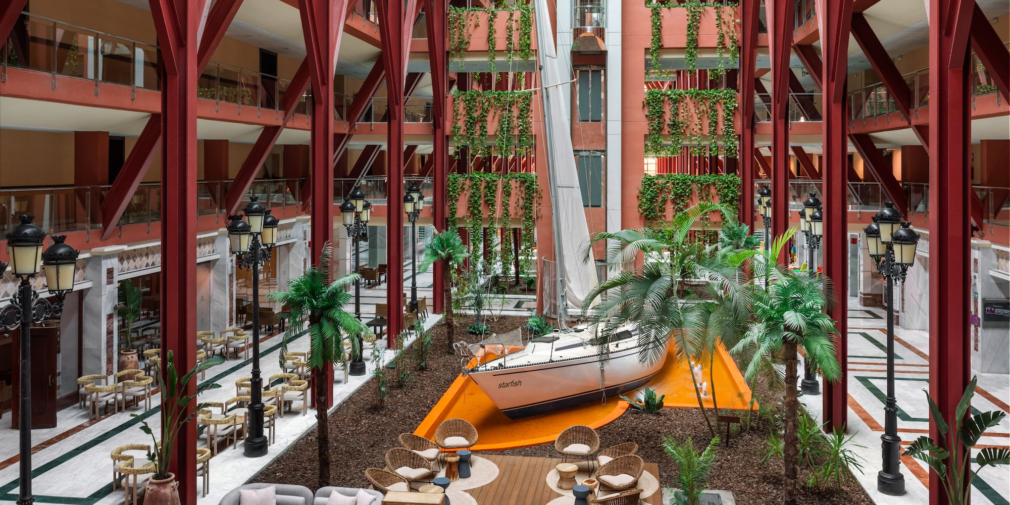 Hotel atrium with large sailboat, red structural beams, vertical gardens, and lounge.
