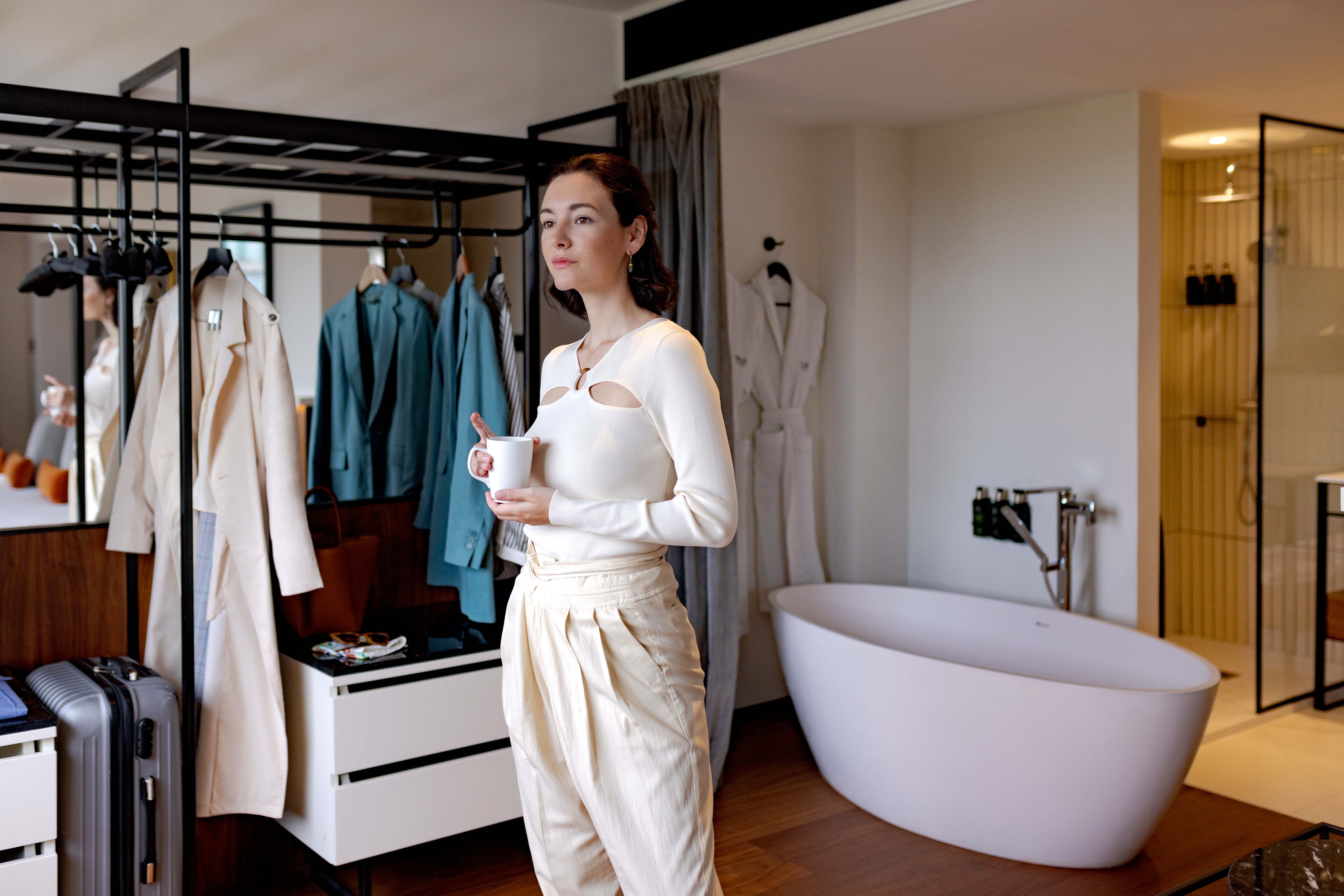 Woman holding mug in modern hotel suite with open closet, freestanding bathtub.