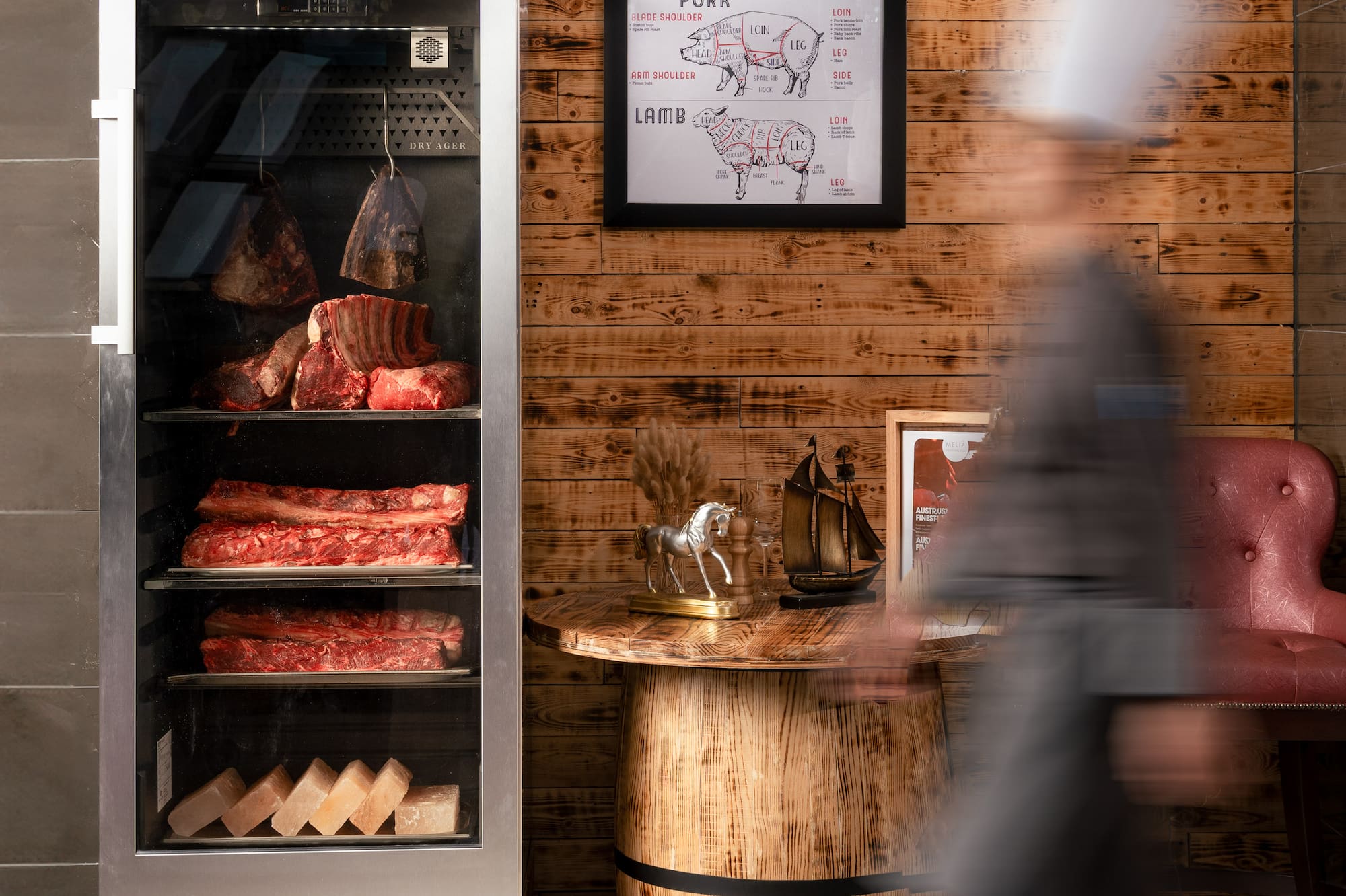 Dry ager displaying beef cuts, meat chart on wood wall, blurred chef walking.