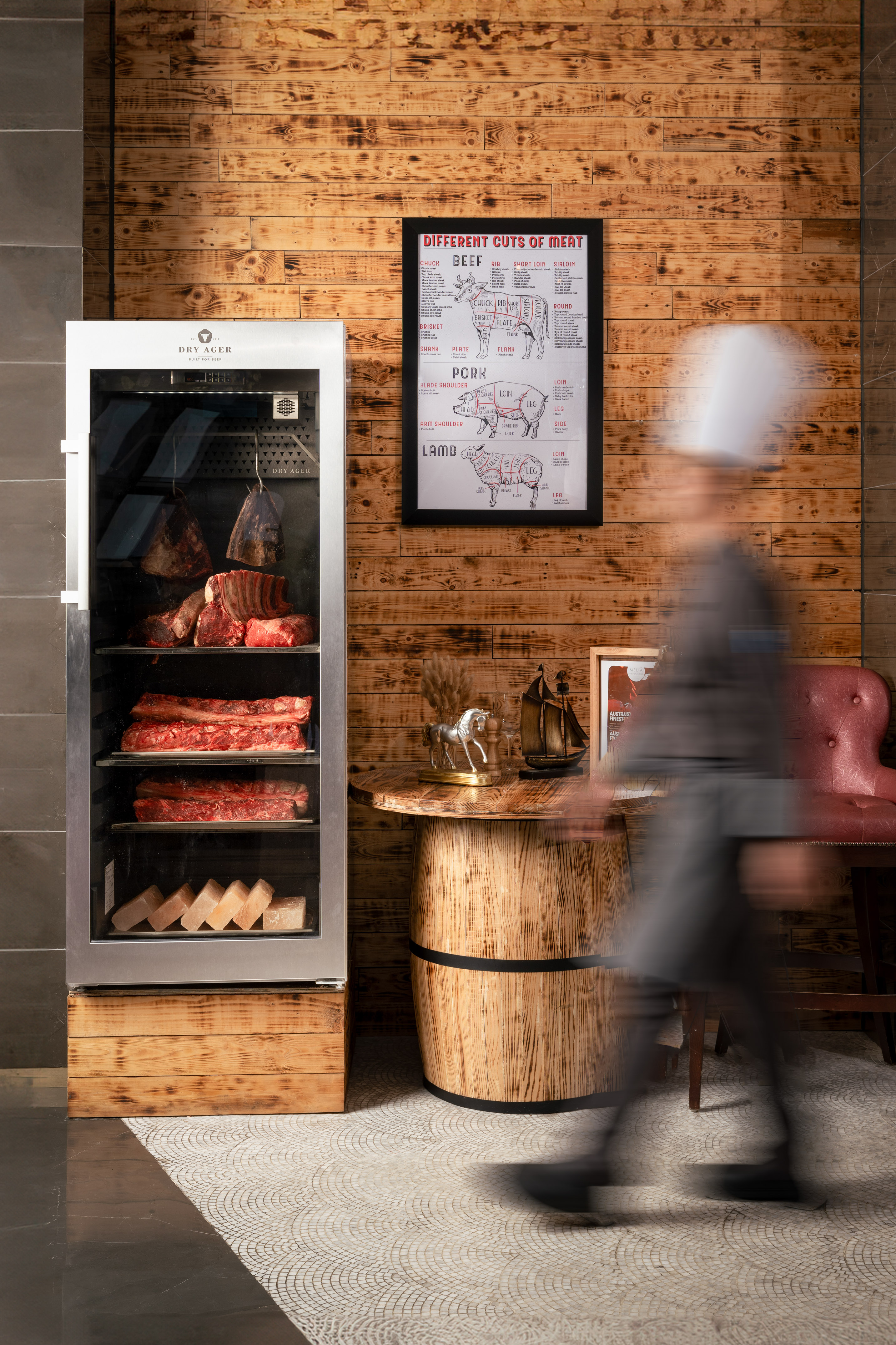 Dry ager displaying beef cuts, meat chart on wood wall, blurred chef walking.