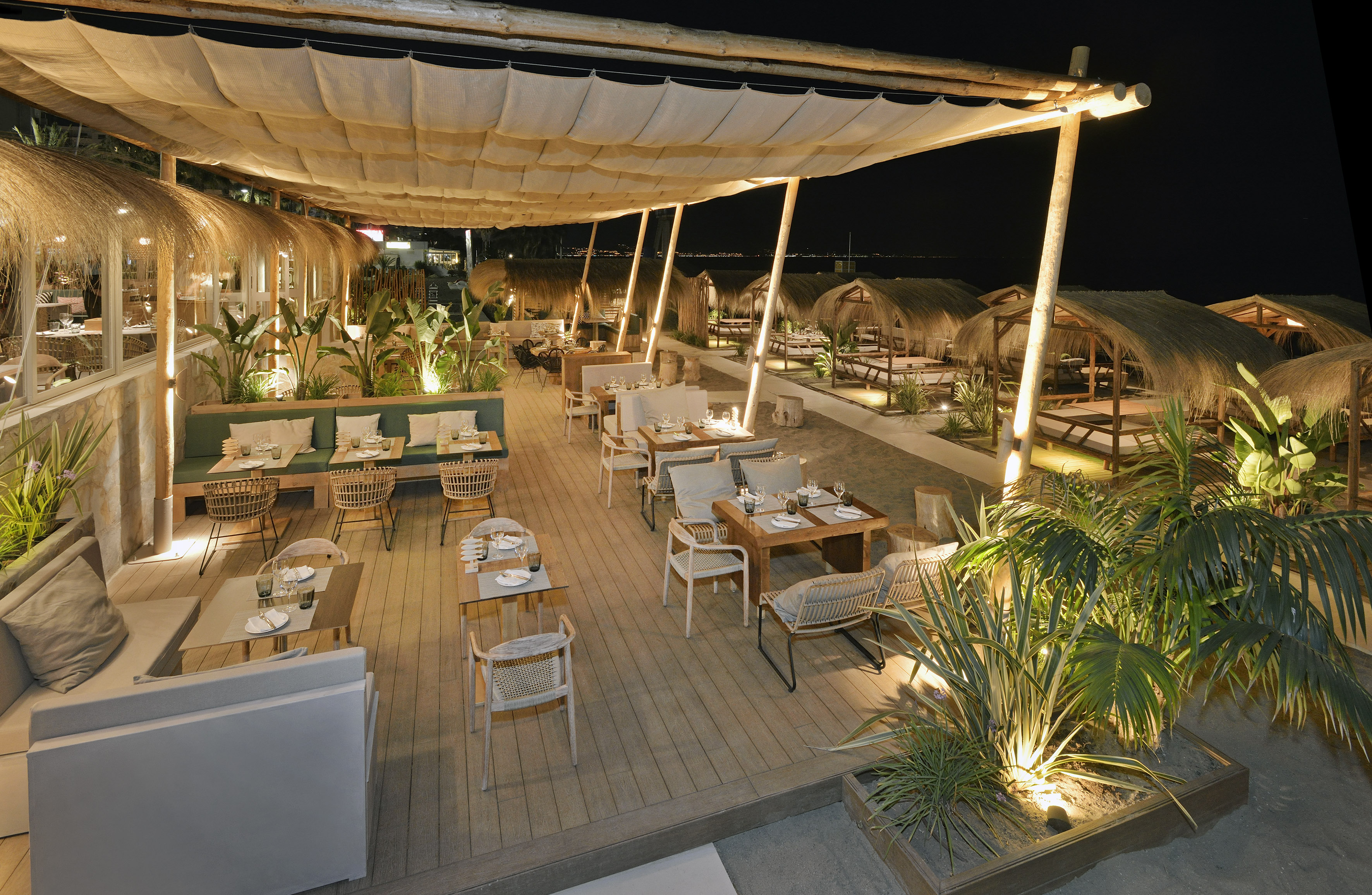 Nighttime beach bar restaurant, elegant outdoor dining area with thatched cabanas.