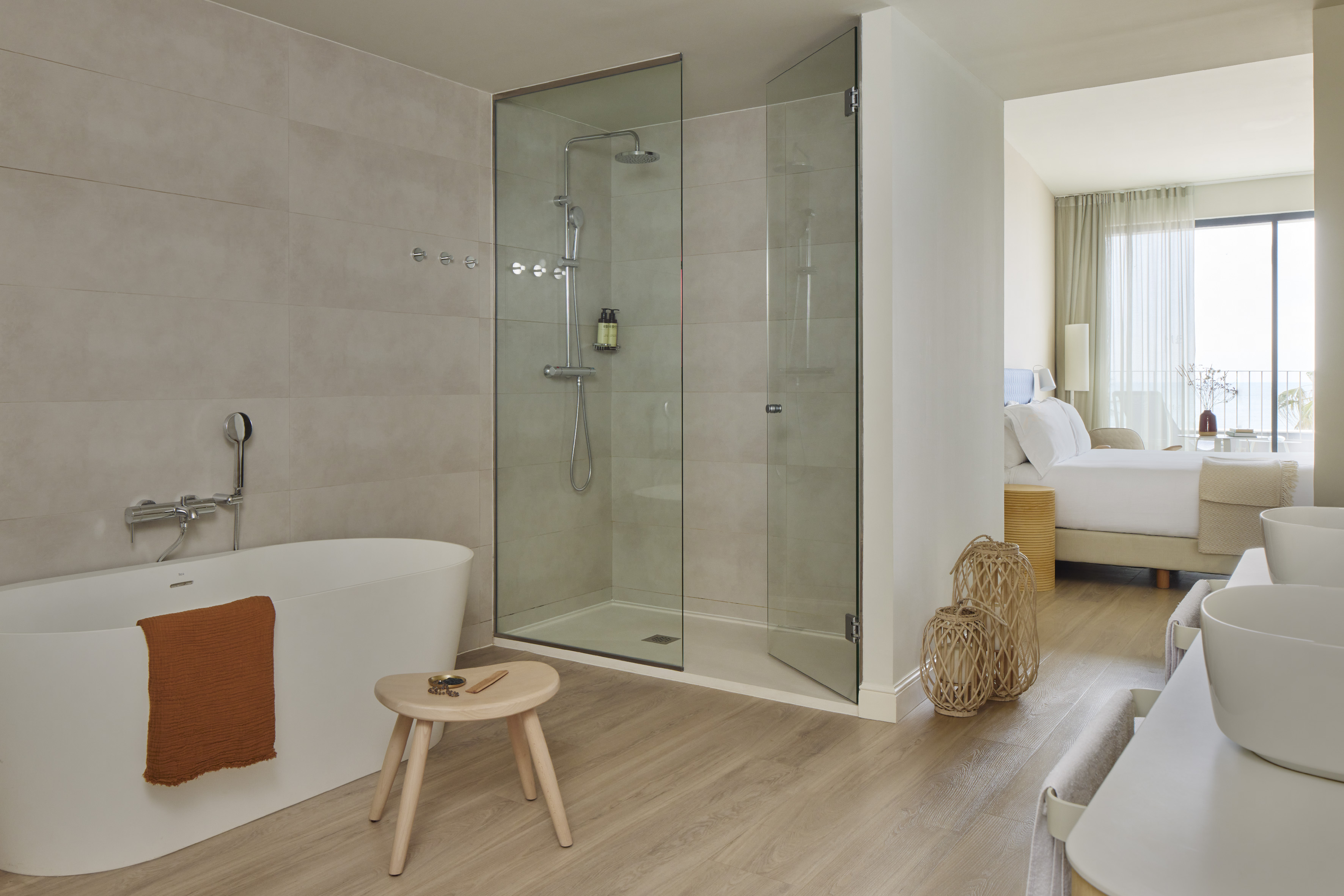 Modern hotel bathroom with freestanding tub, glass shower, and bedroom view.