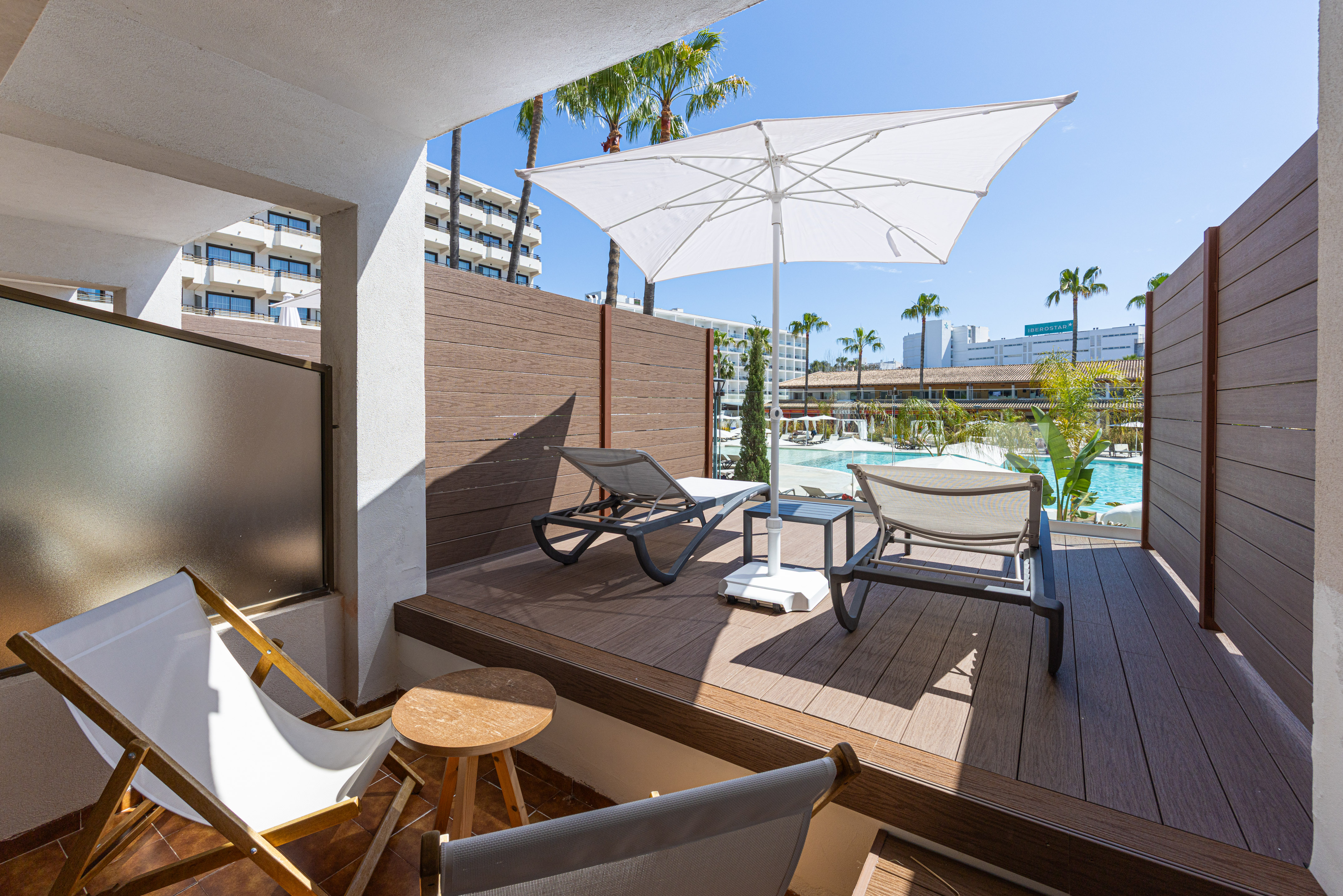 Hotel balcony with pool view, lounge chairs, umbrella, and palm trees.