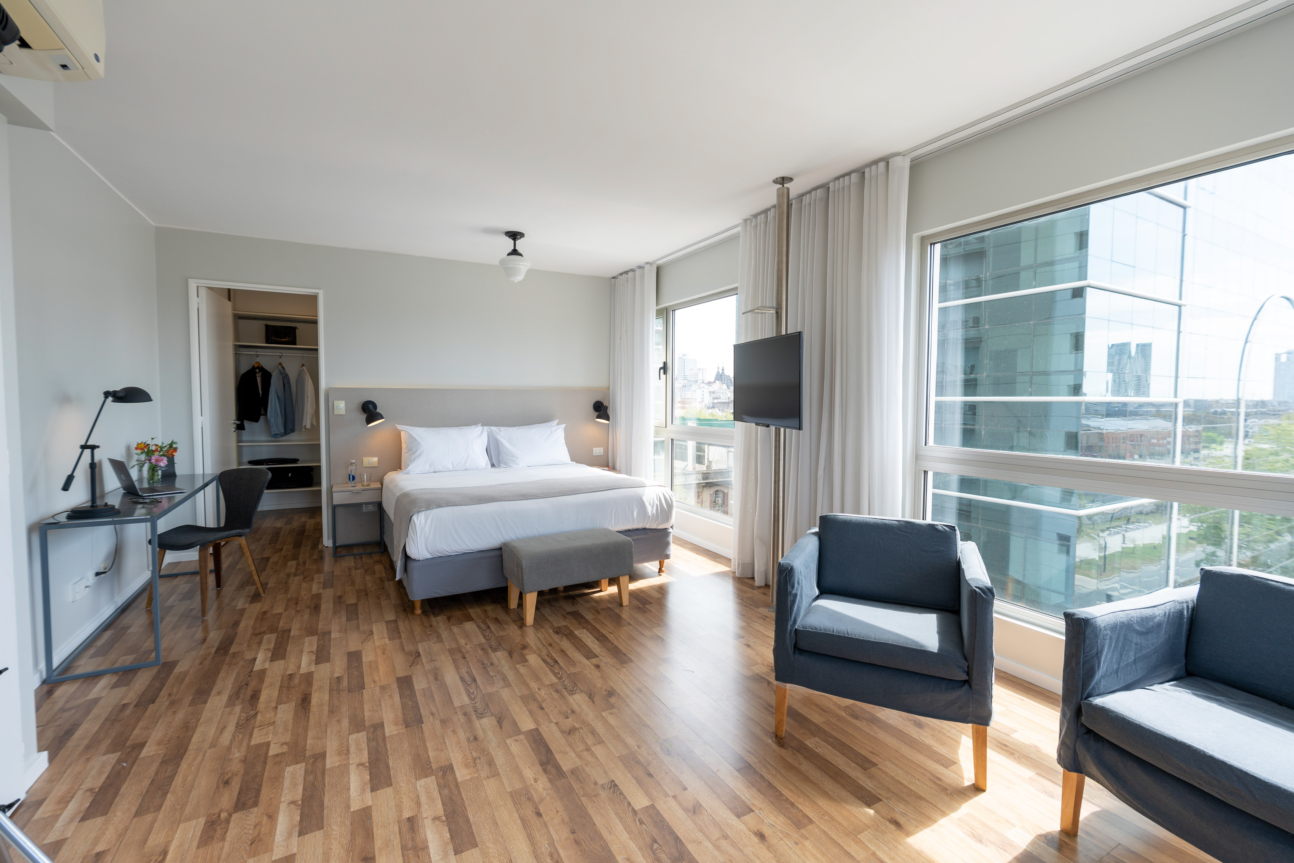 Modern hotel room with bed, desk, wood floor, and large city view windows.