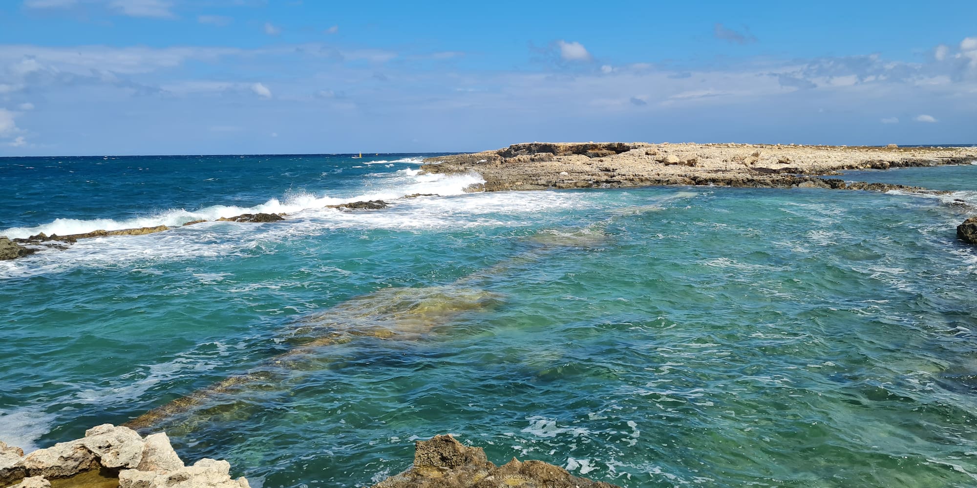 Rocky turquoise coastline with crashing waves under a bright blue sky.