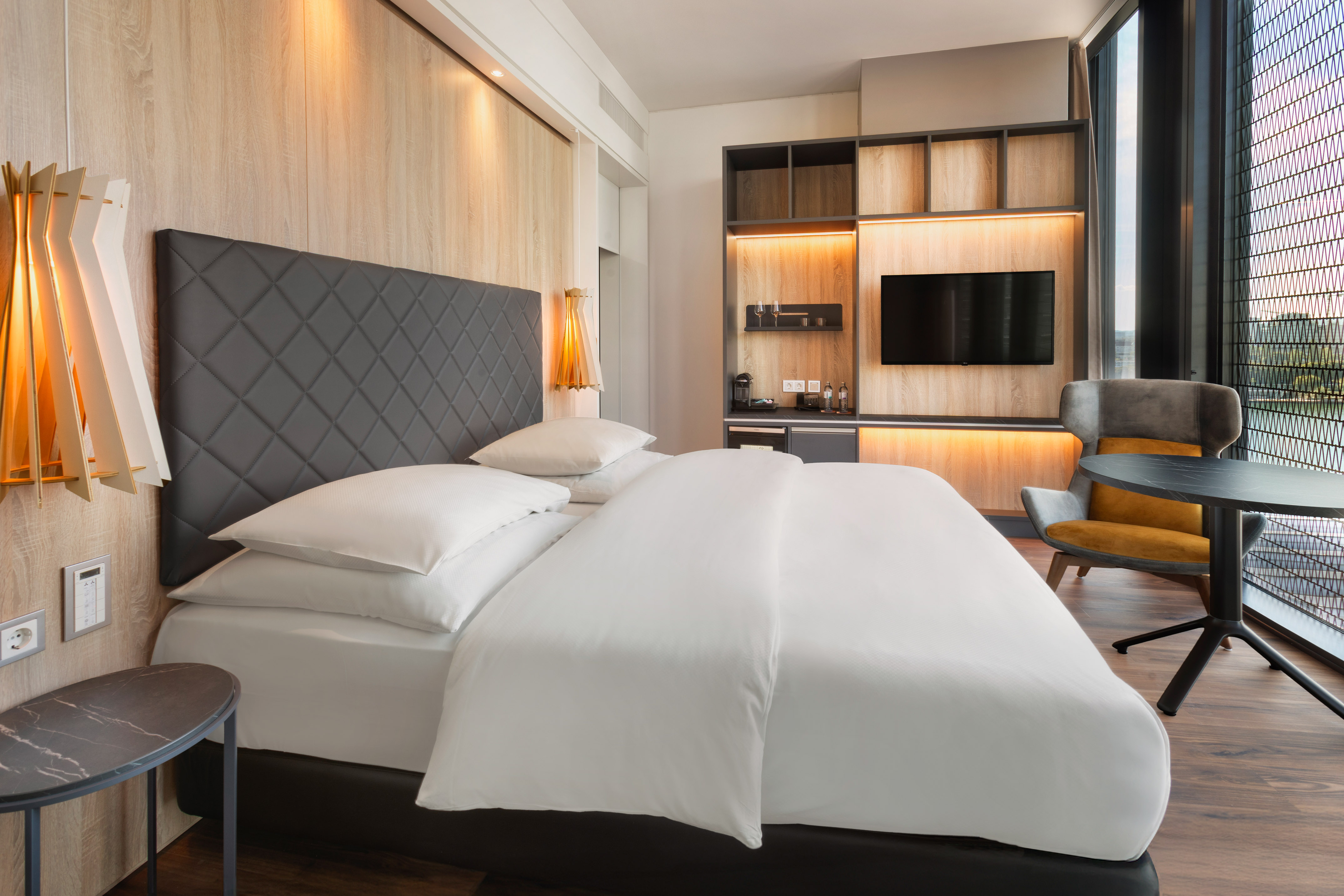 Modern hotel room with large bed, wood wall, TV, comfortable armchair, and window.