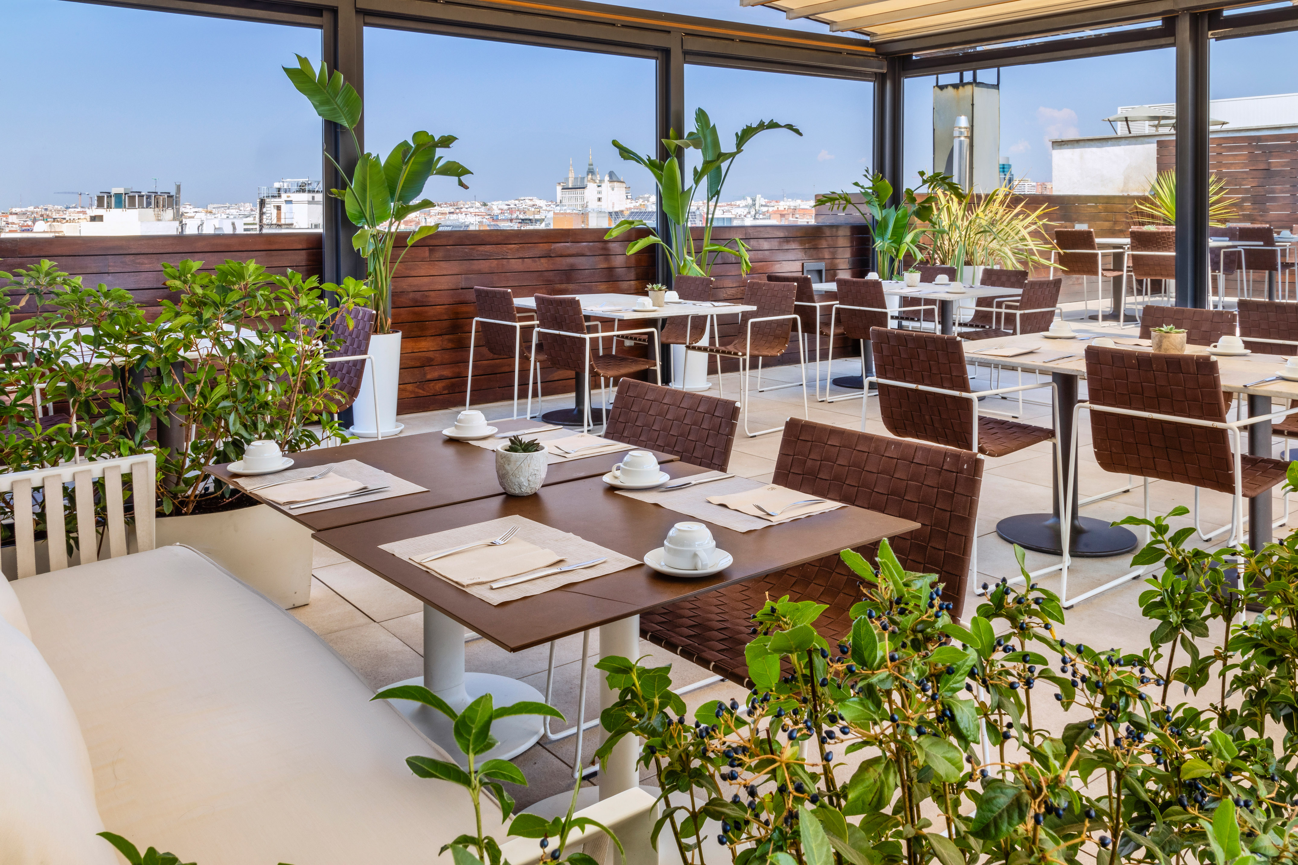 Outdoor rooftop dining terrace with city views, set tables, and lush green plants.
