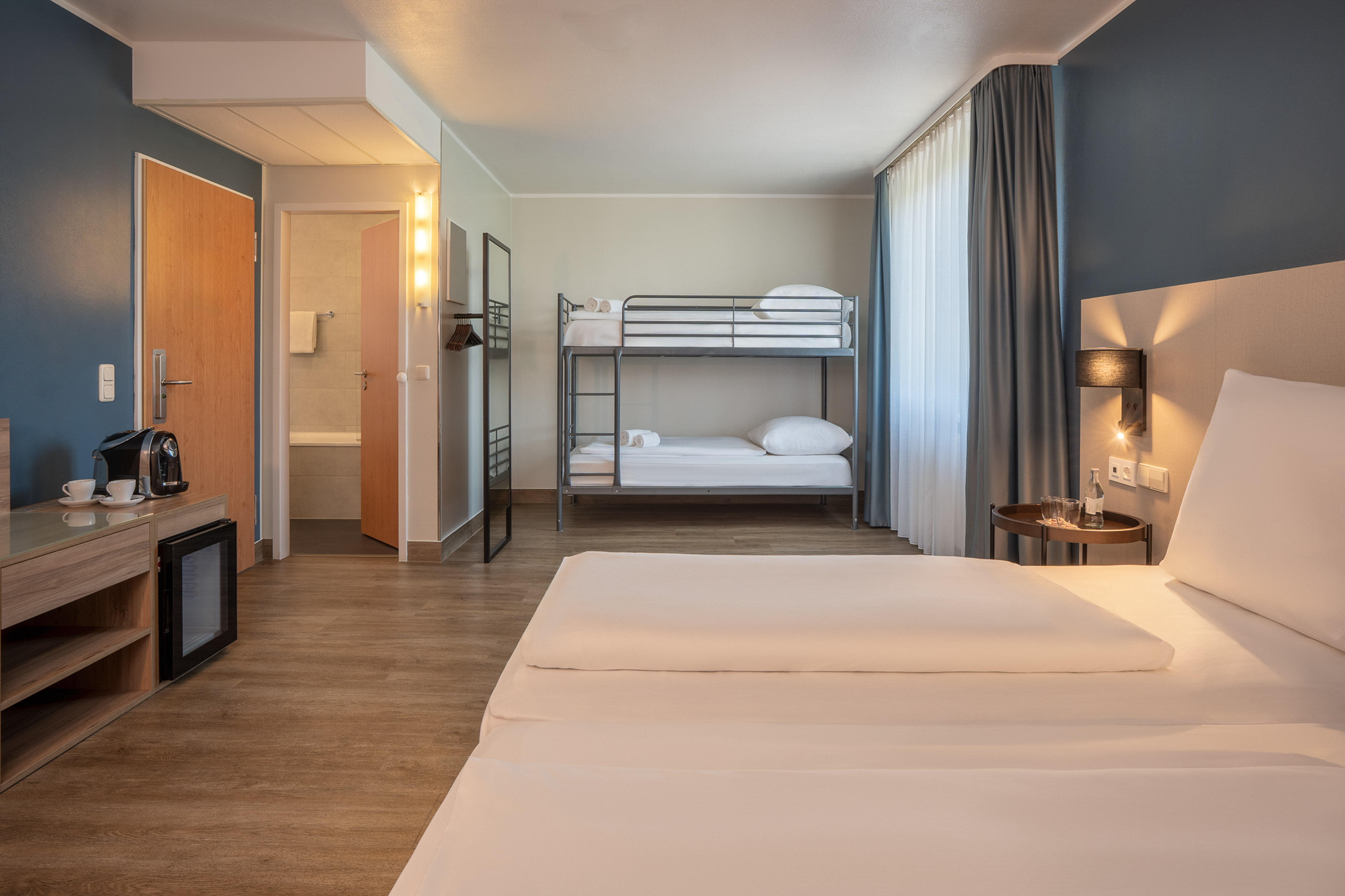 Modern hotel family room with queen bed and metal bunk beds.