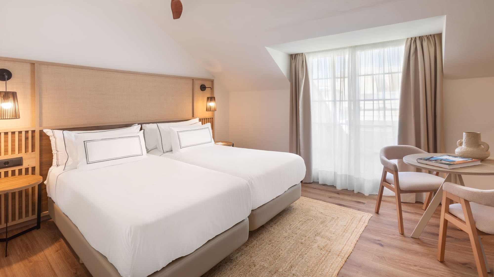 Modern hotel room with twin beds, ceiling fan, woven headboard, and seating area.