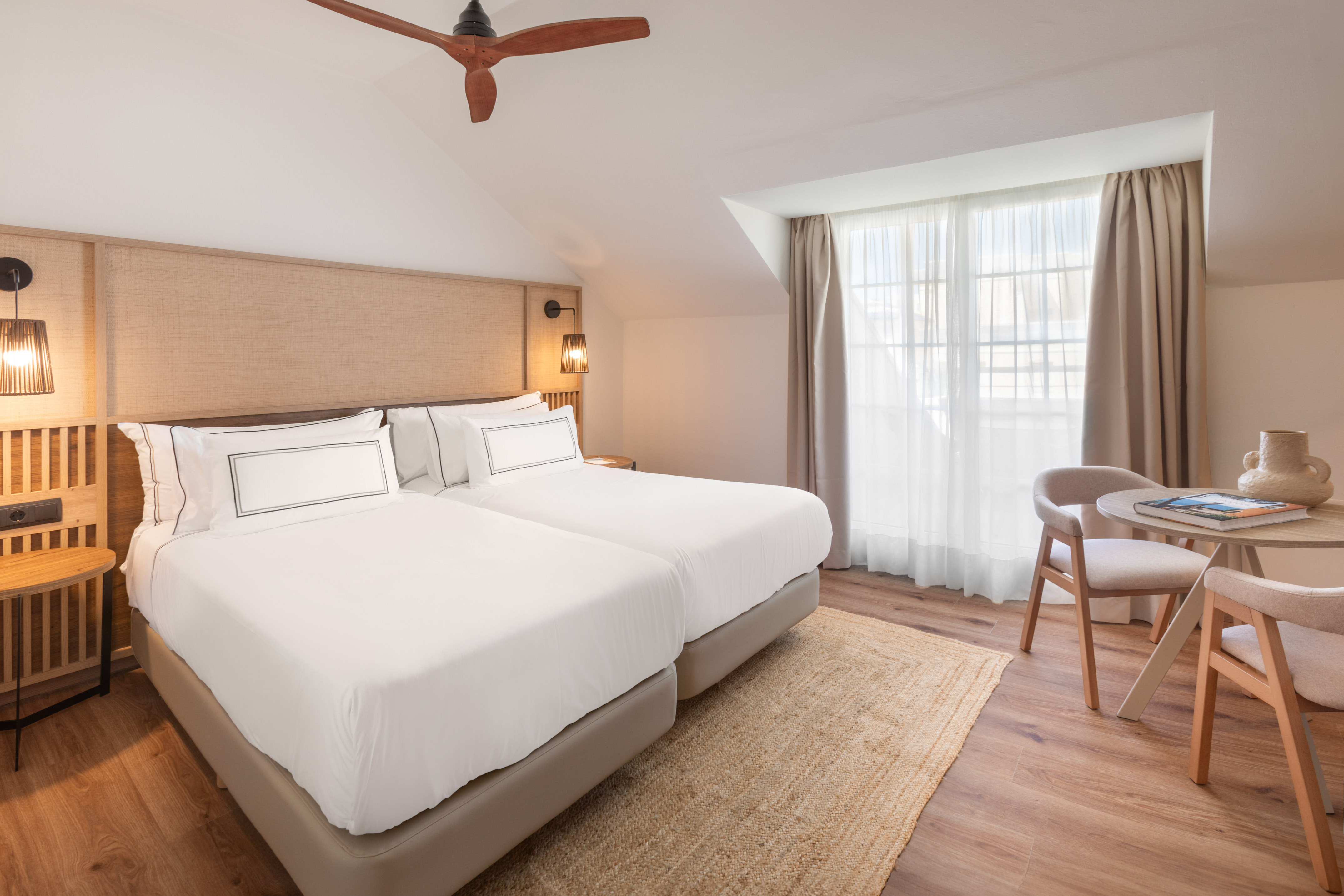Modern hotel room with twin beds, ceiling fan, woven headboard, and seating area.