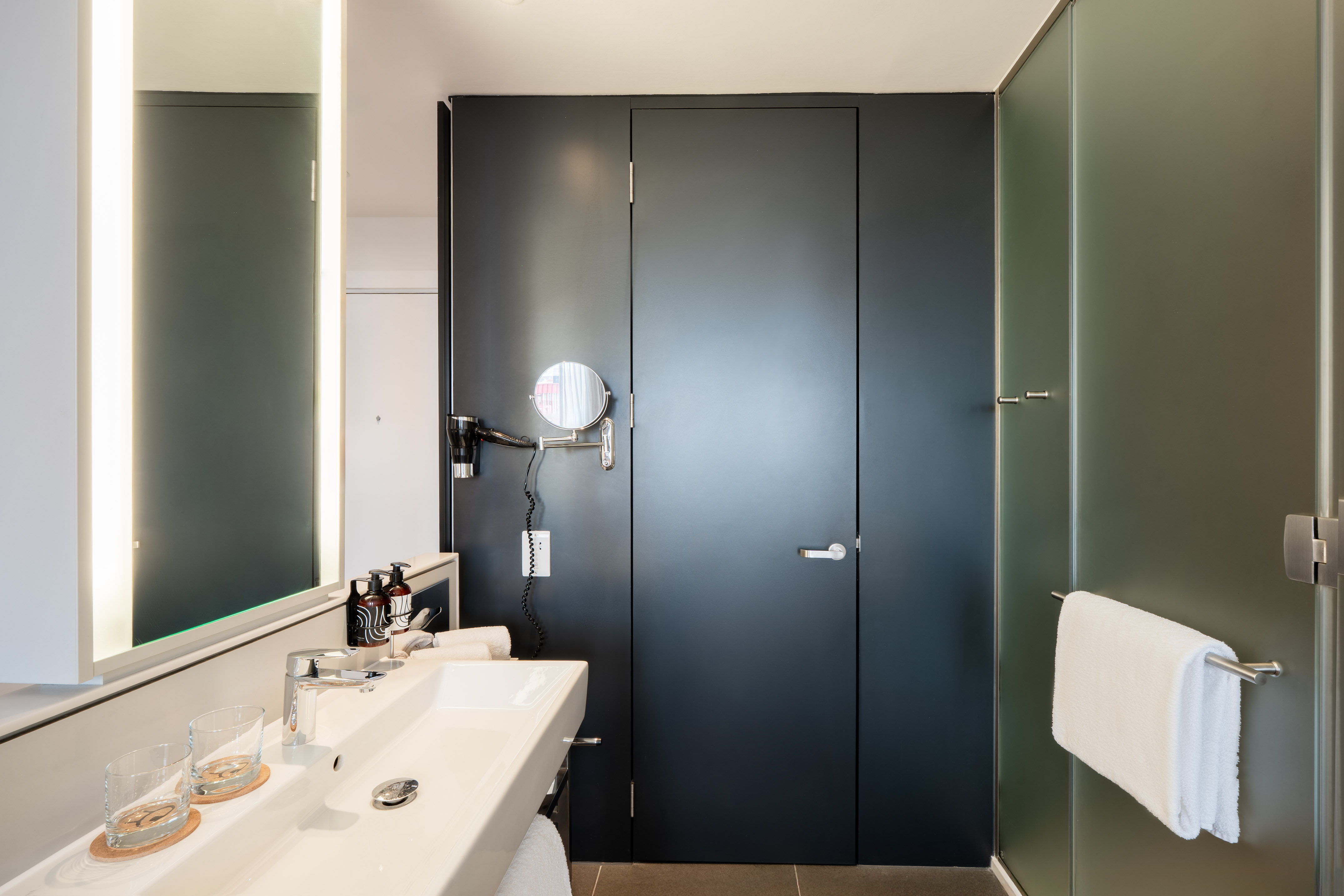Modern hotel bathroom with white vanity, lighted mirror, and glass shower.