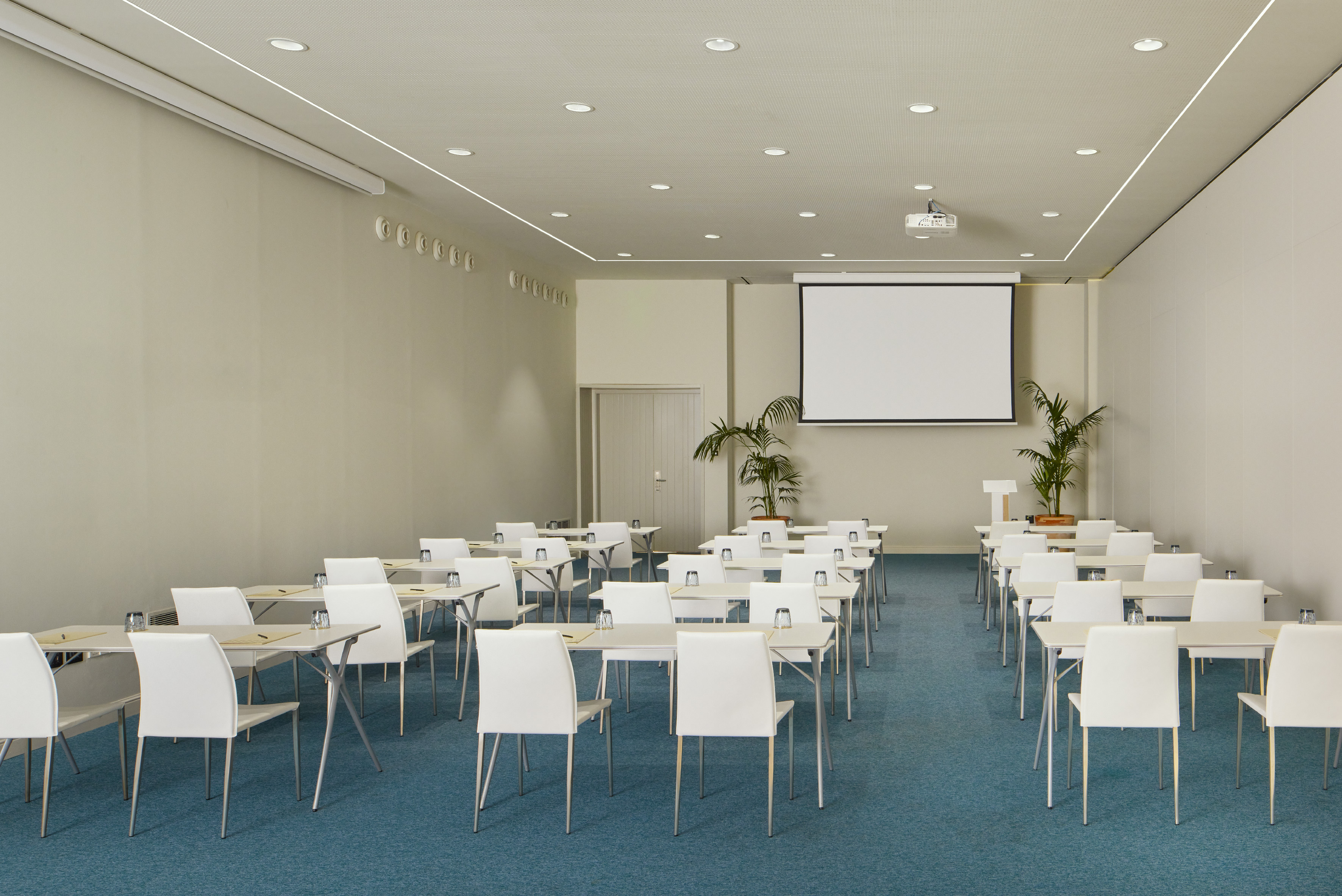 Clean conference room with white tables, chairs, projector screen, and blue carpet.