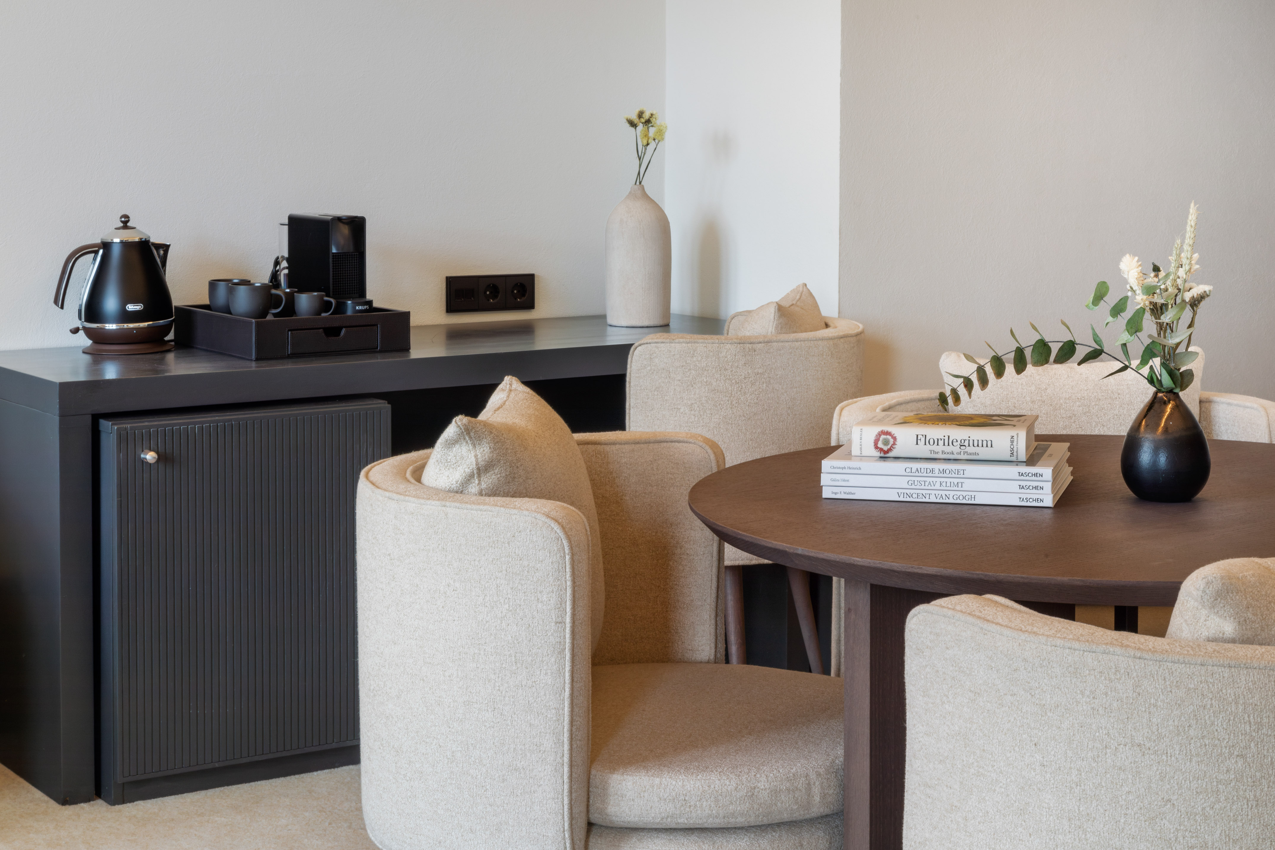 Modern hotel room with coffee station, minibar, round table, and upholstered chairs.