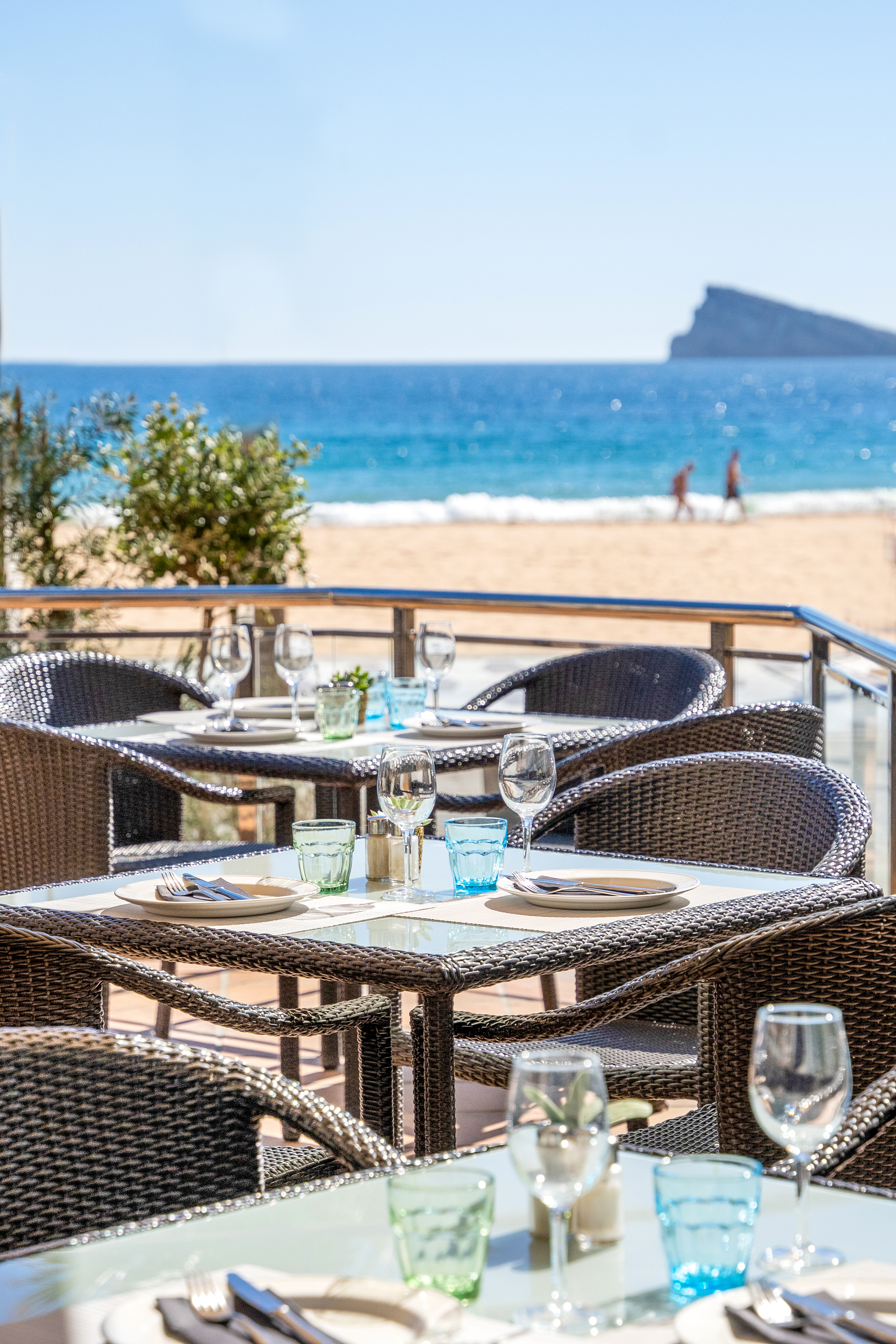 Dining tables on a coastal terrace overlooking a blue sea and sandy beach.