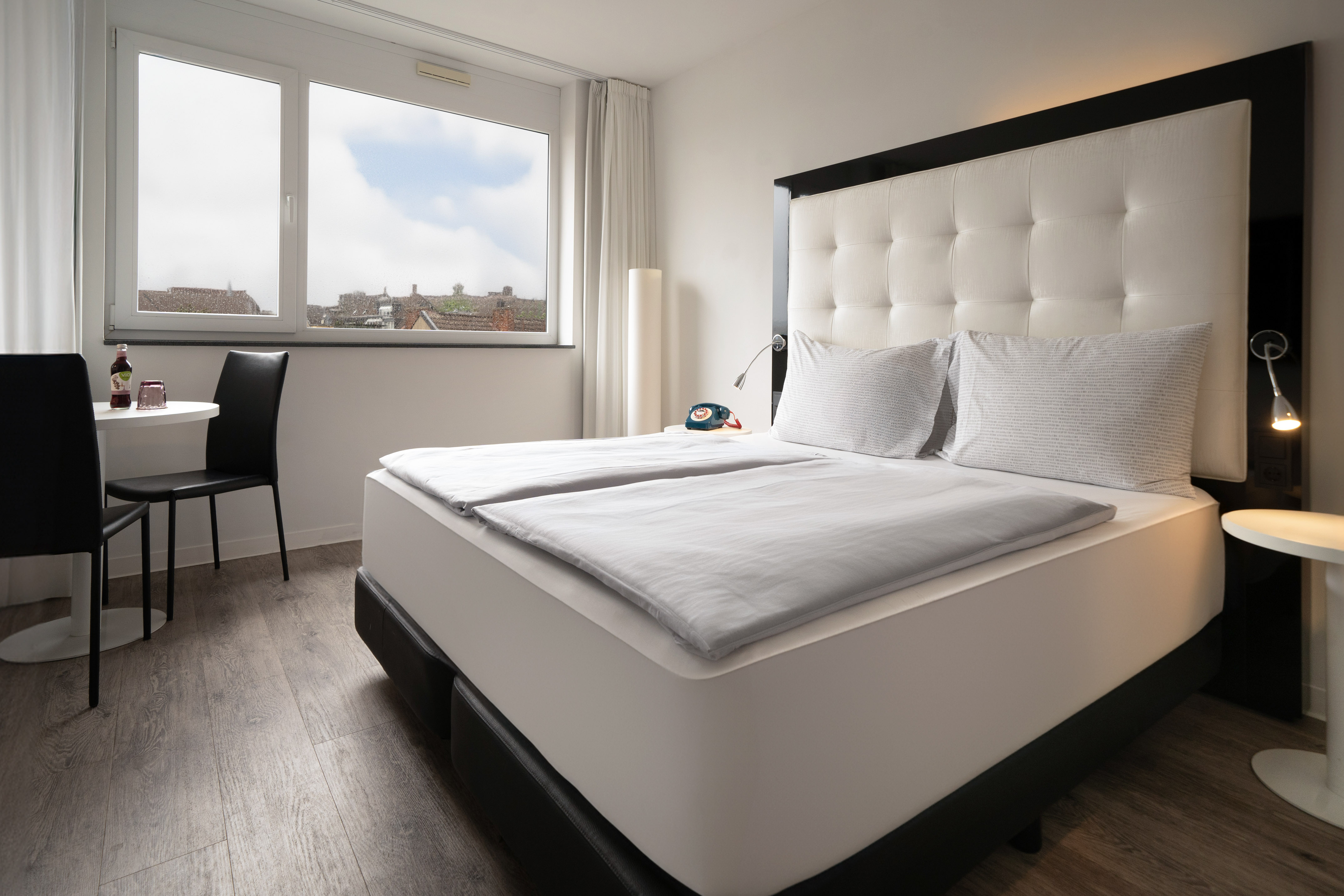 Modern hotel room with a double bed, large window city view, and small table.