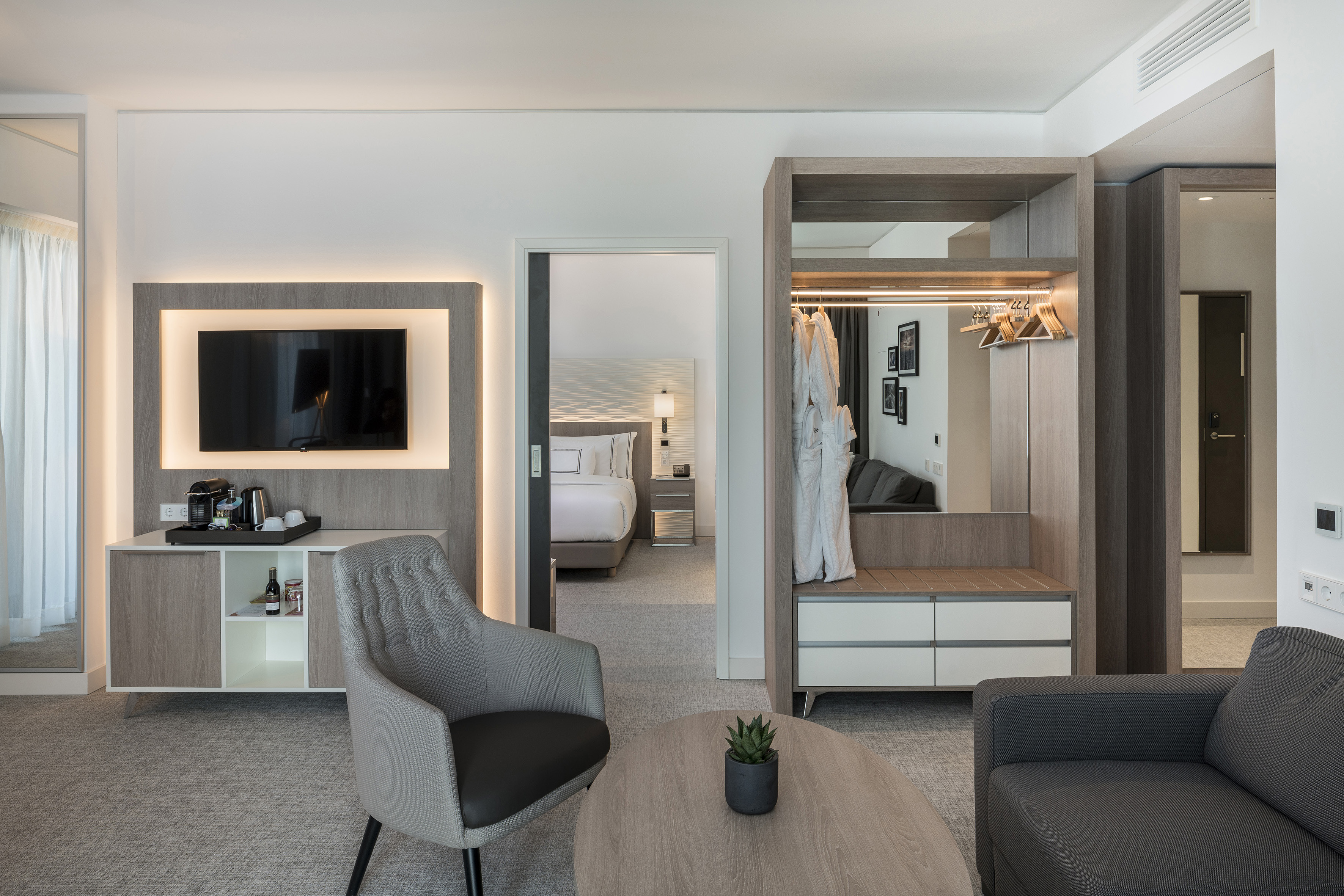 Modern hotel suite with lounge area, backlit TV, open wardrobe, and bedroom.