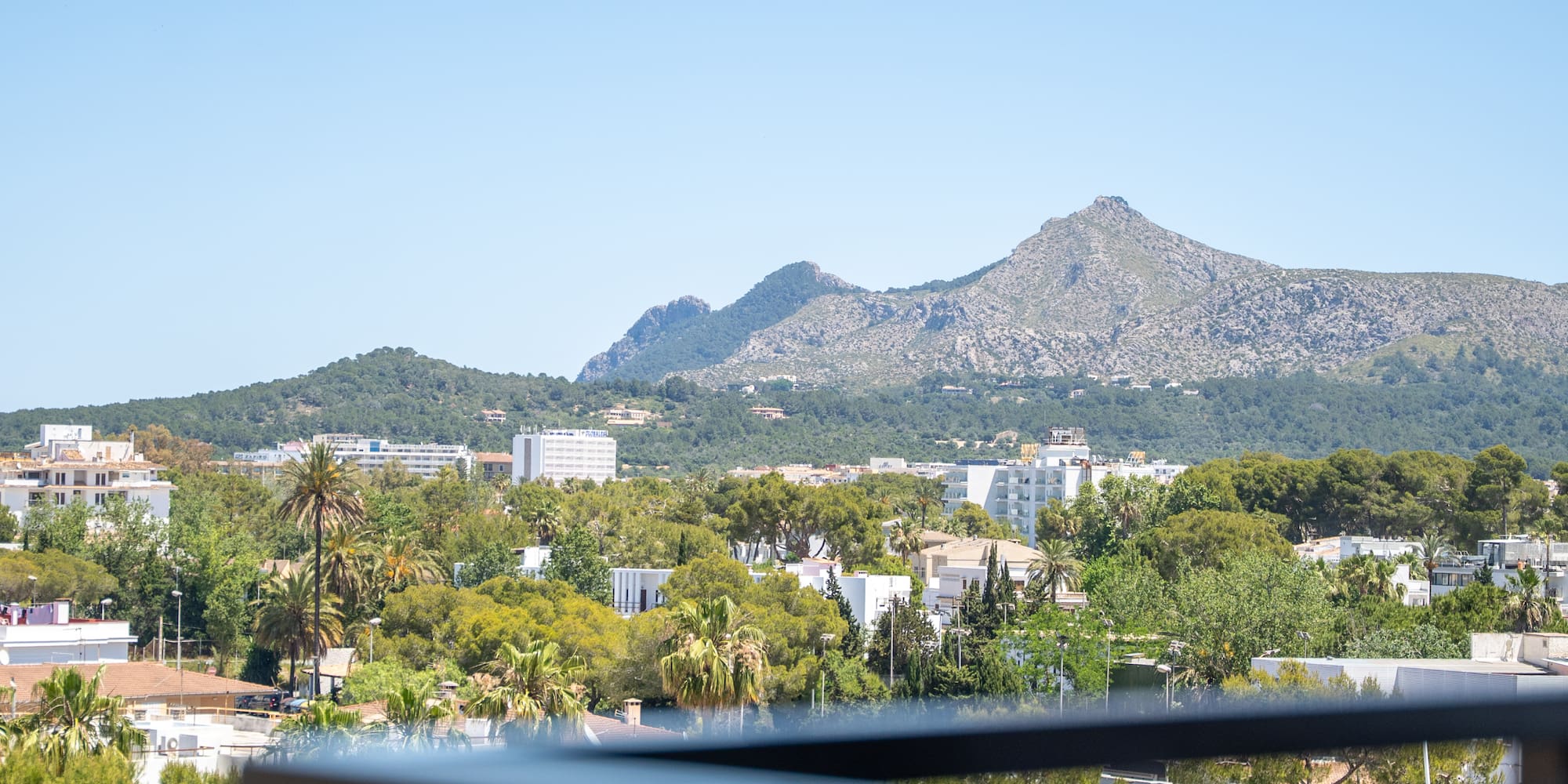 Scenic balcony view of a sunny town, green trees, and distant mountains.