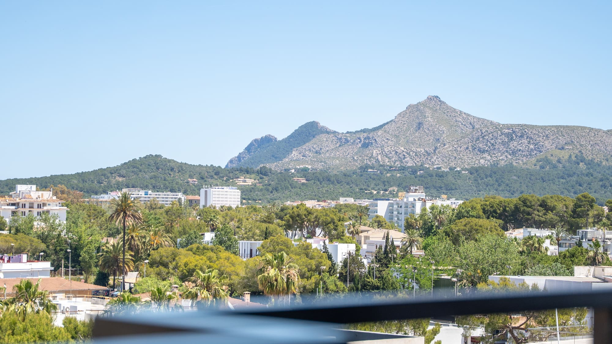 Scenic balcony view of a sunny town, green trees, and distant mountains.
