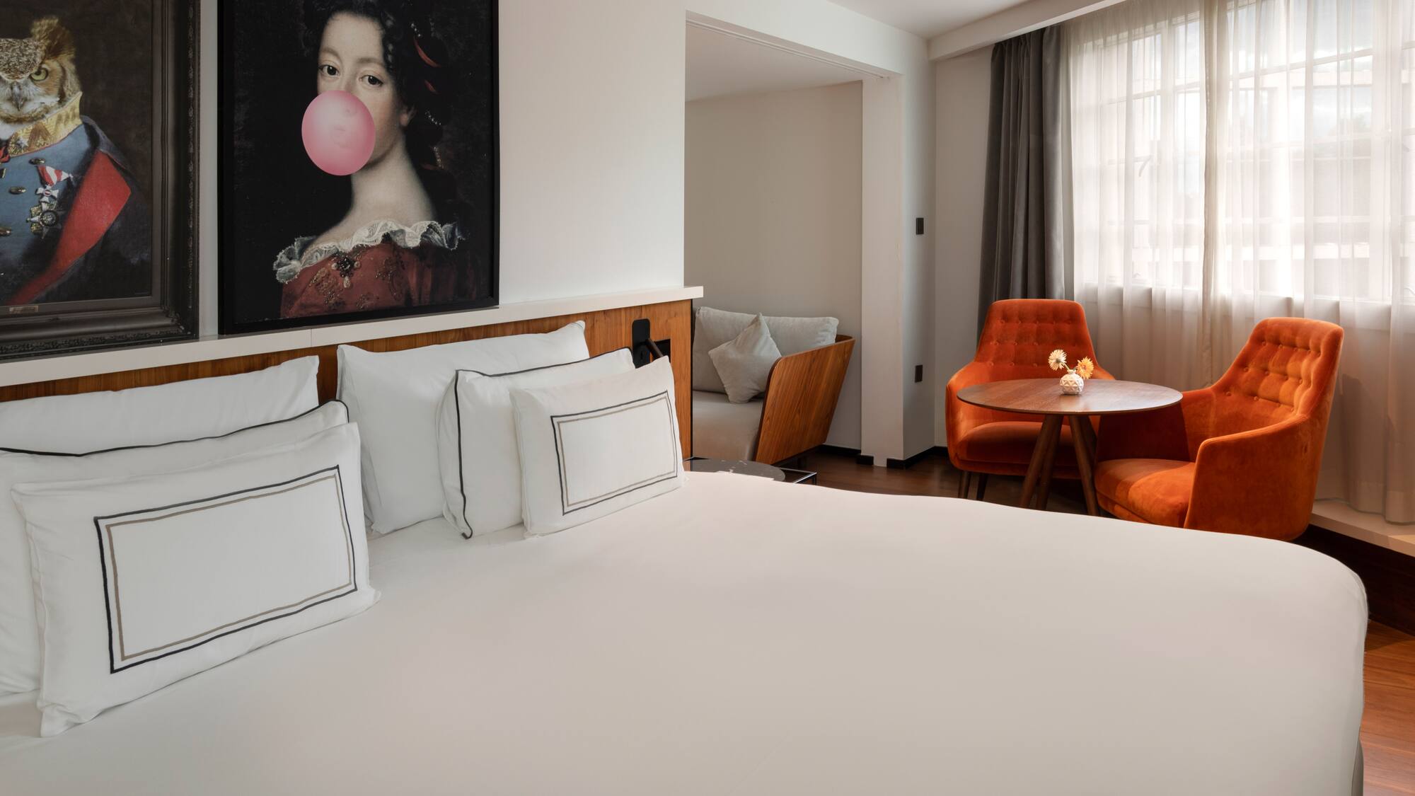 Hotel room with white bed, bubblegum painting, and orange tufted armchairs.
