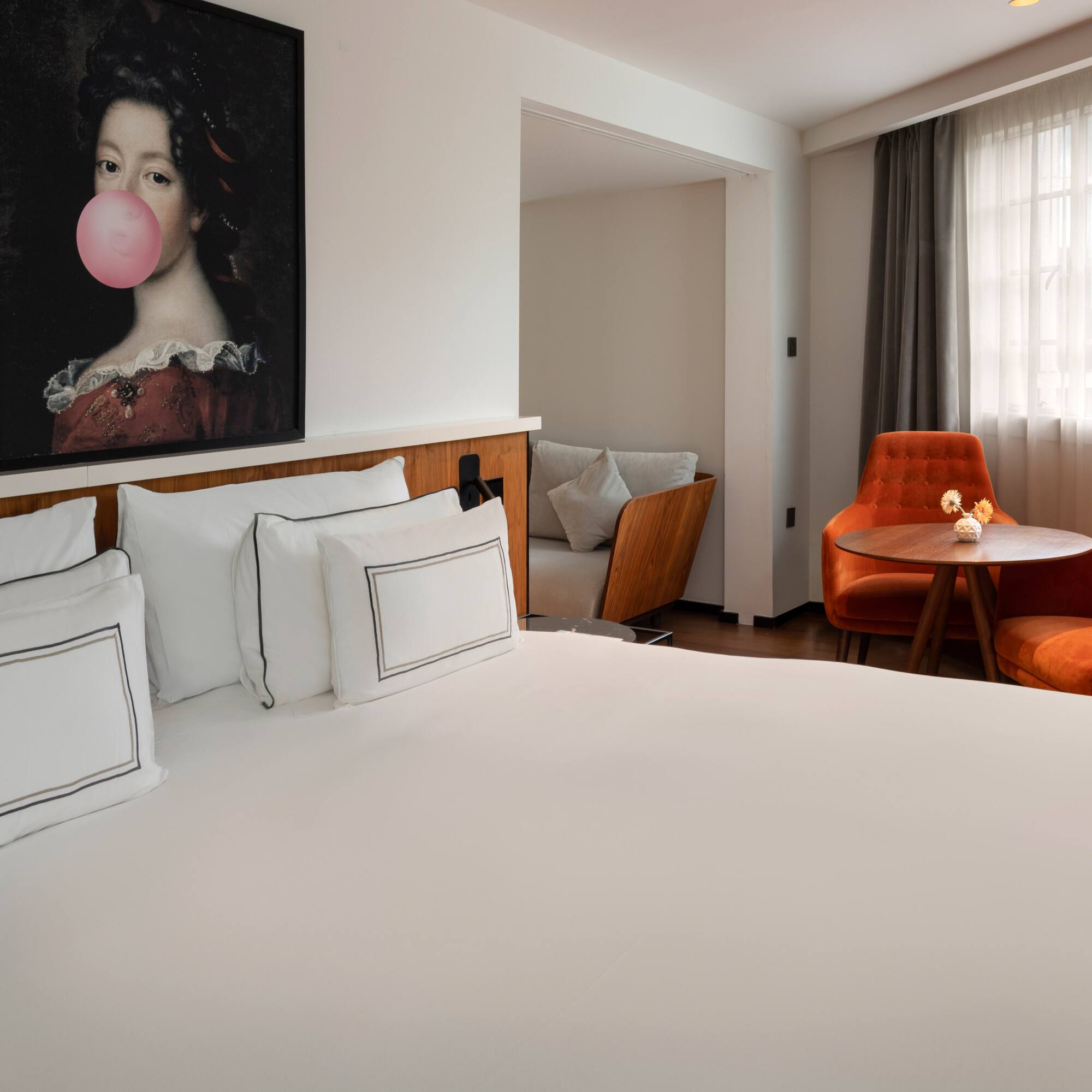 Hotel room with white bed, bubblegum painting, and orange tufted armchairs.