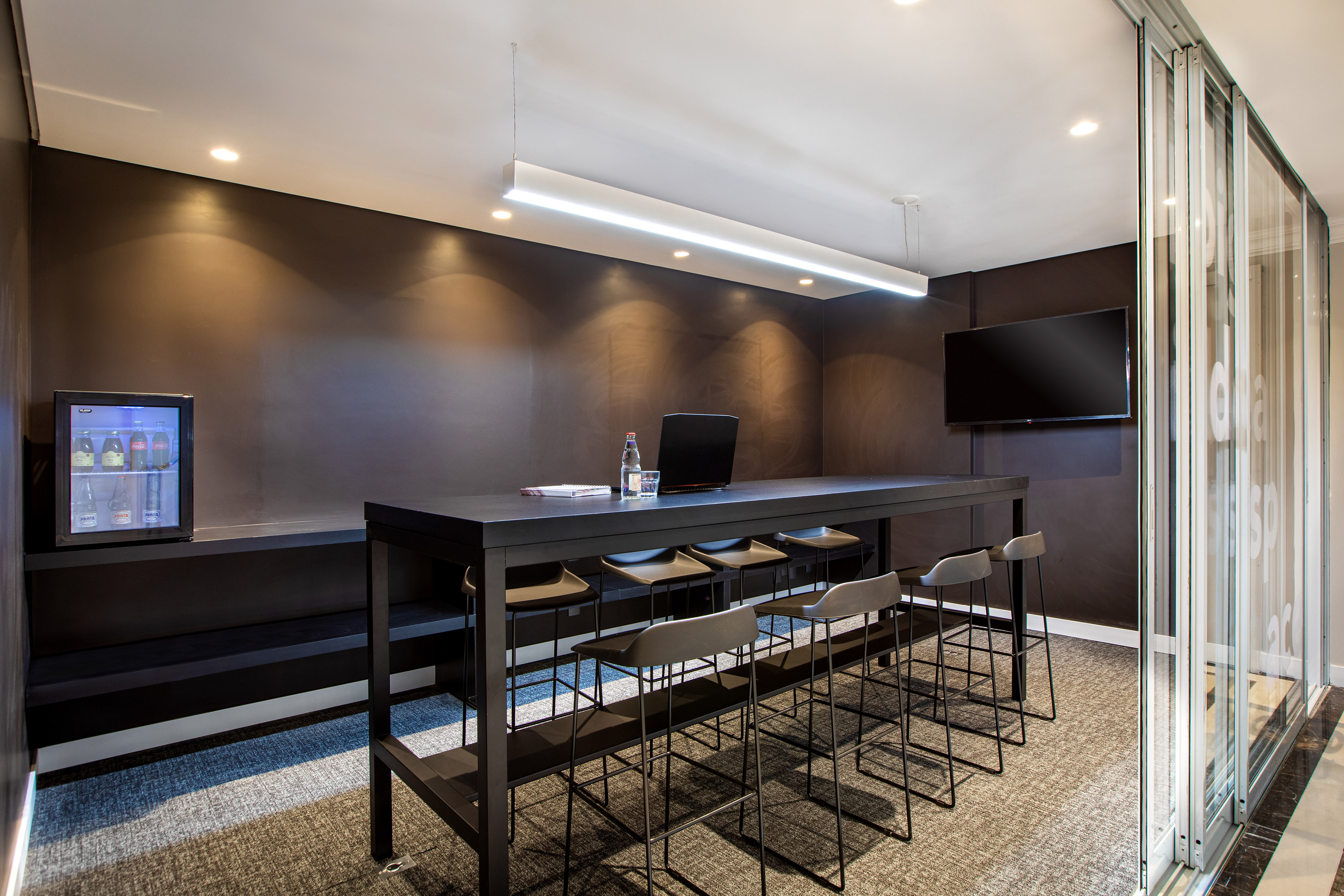 Modern office lounge with high-top table, bar stools, TV, and mini-fridge.
