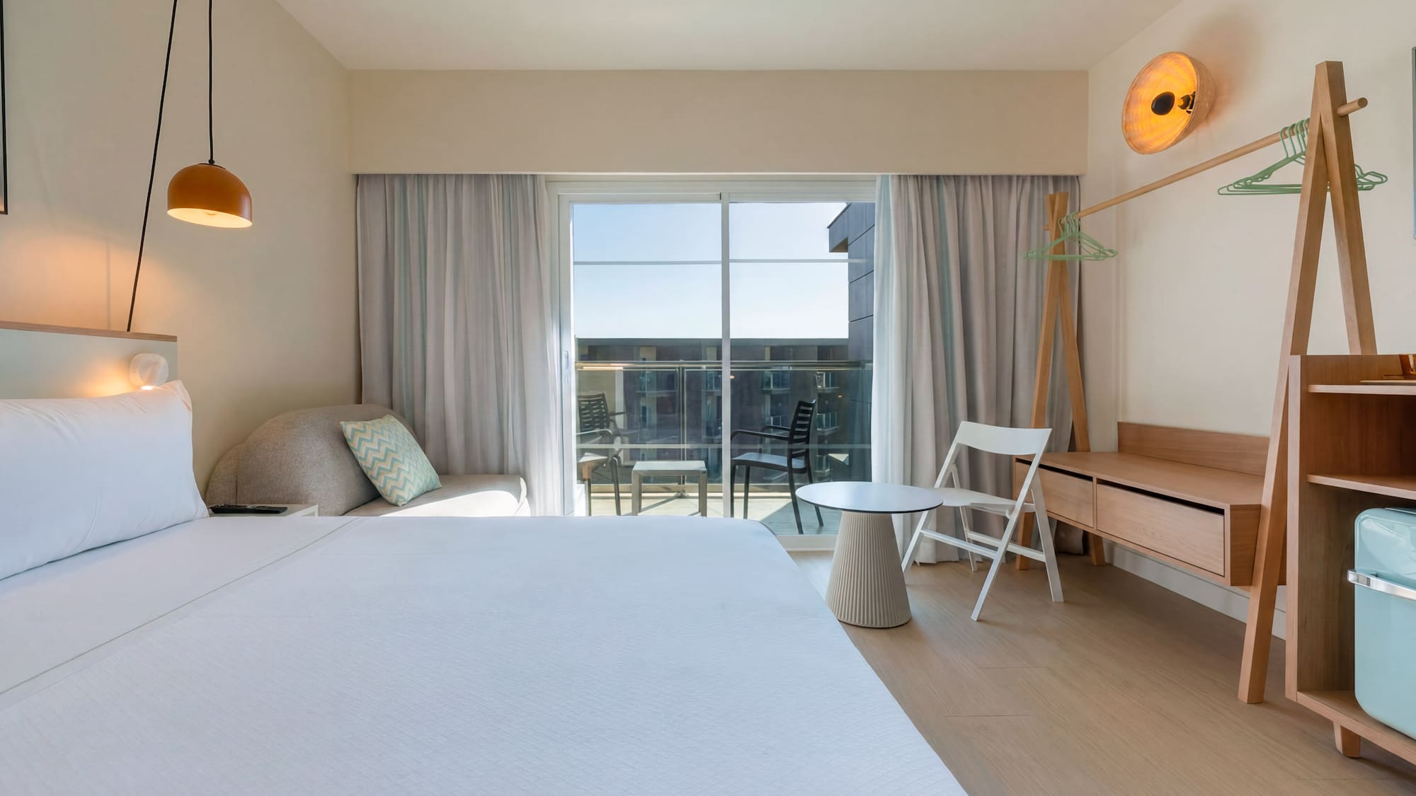 Bright hotel room with large bed, balcony, and modern light wood furniture.