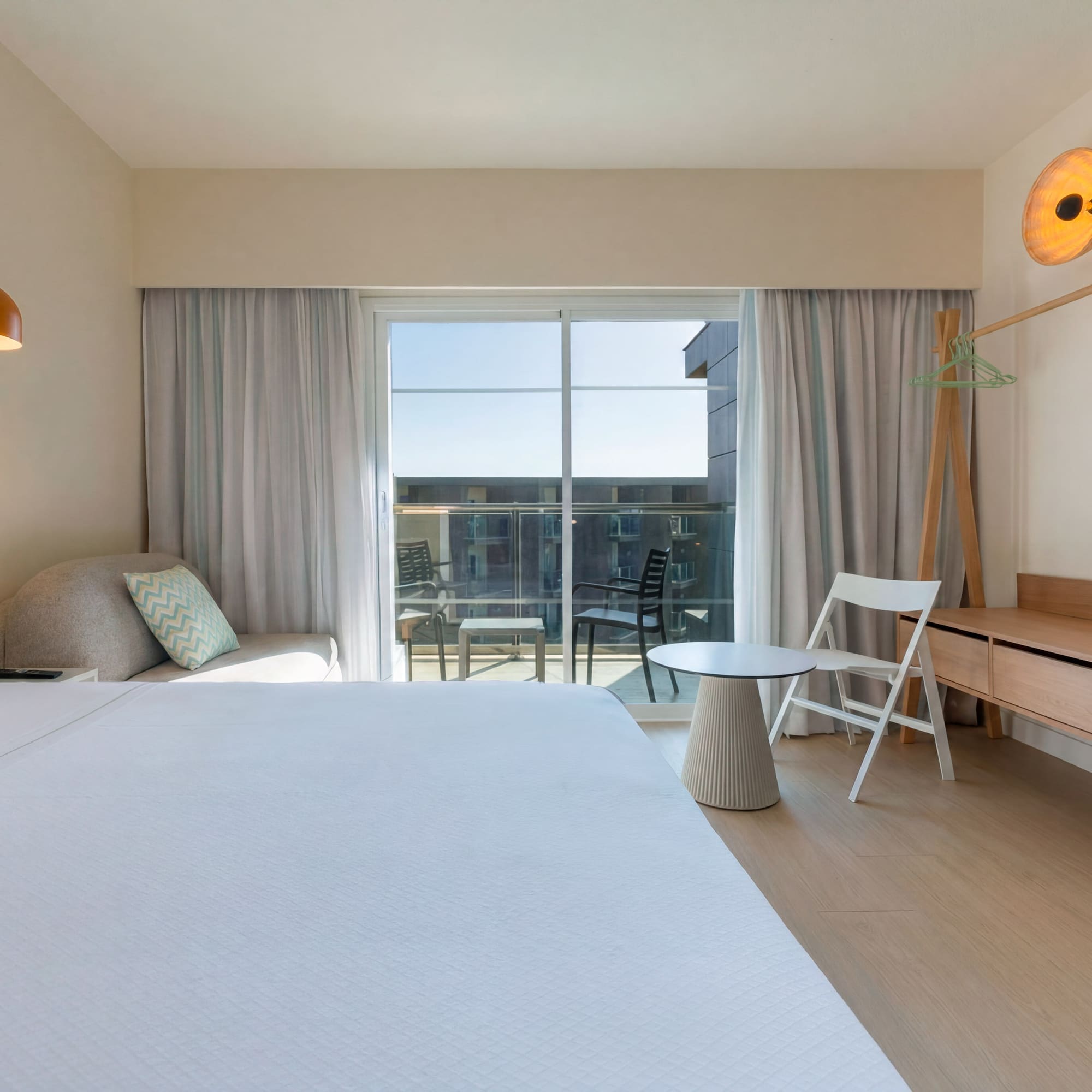 Bright hotel room with large bed, balcony, and modern light wood furniture.