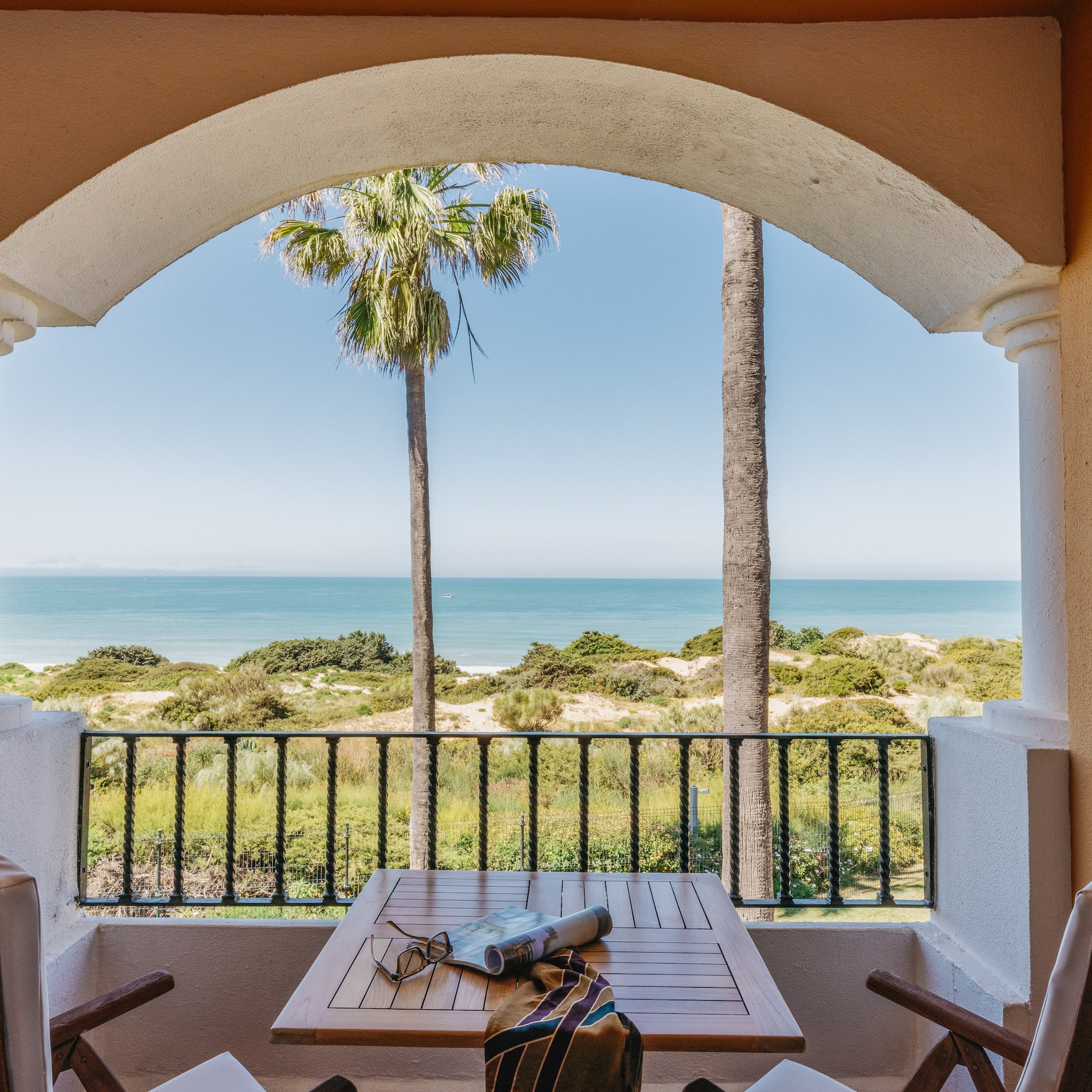 Resort balcony with ocean view, palm trees, beach dunes, table, and chairs.