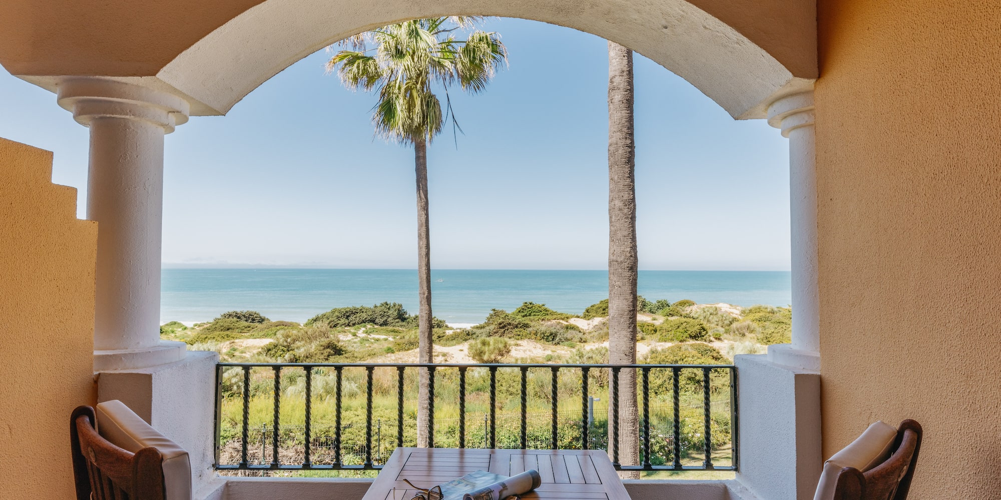 Resort balcony with ocean view, palm trees, beach dunes, table, and chairs.