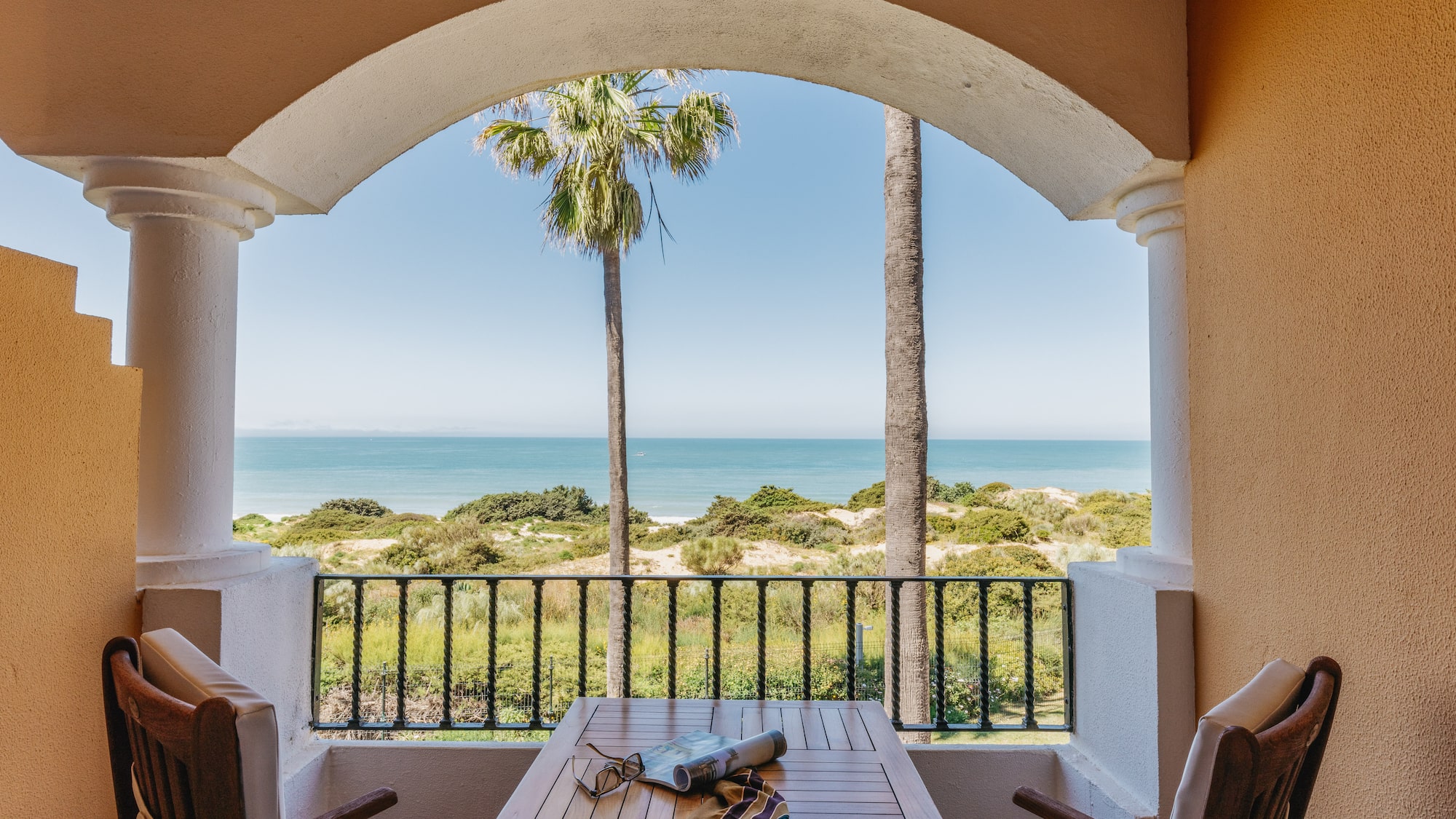 Resort balcony with ocean view, palm trees, beach dunes, table, and chairs.