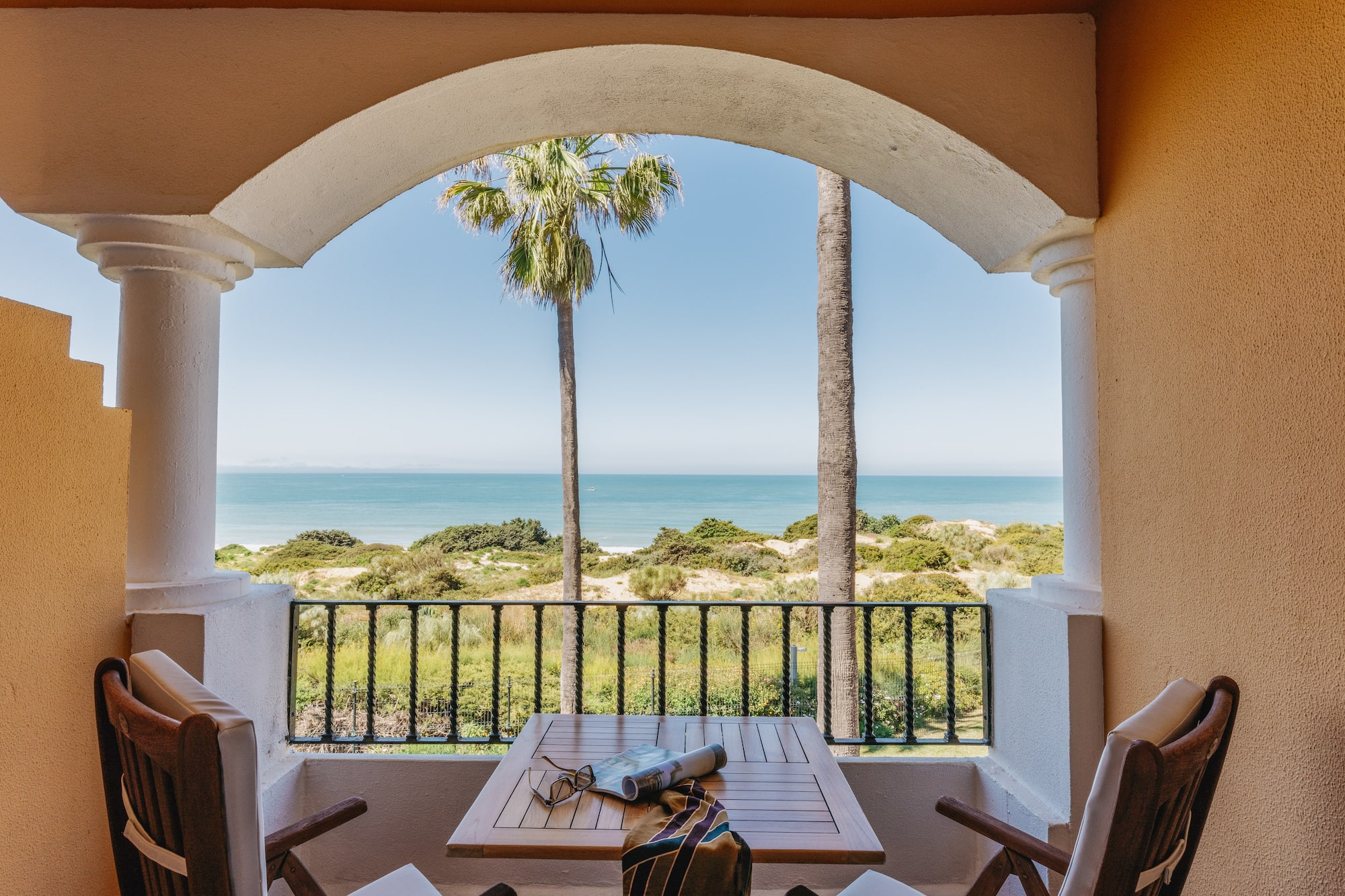 Resort balcony with ocean view, palm trees, beach dunes, table, and chairs.