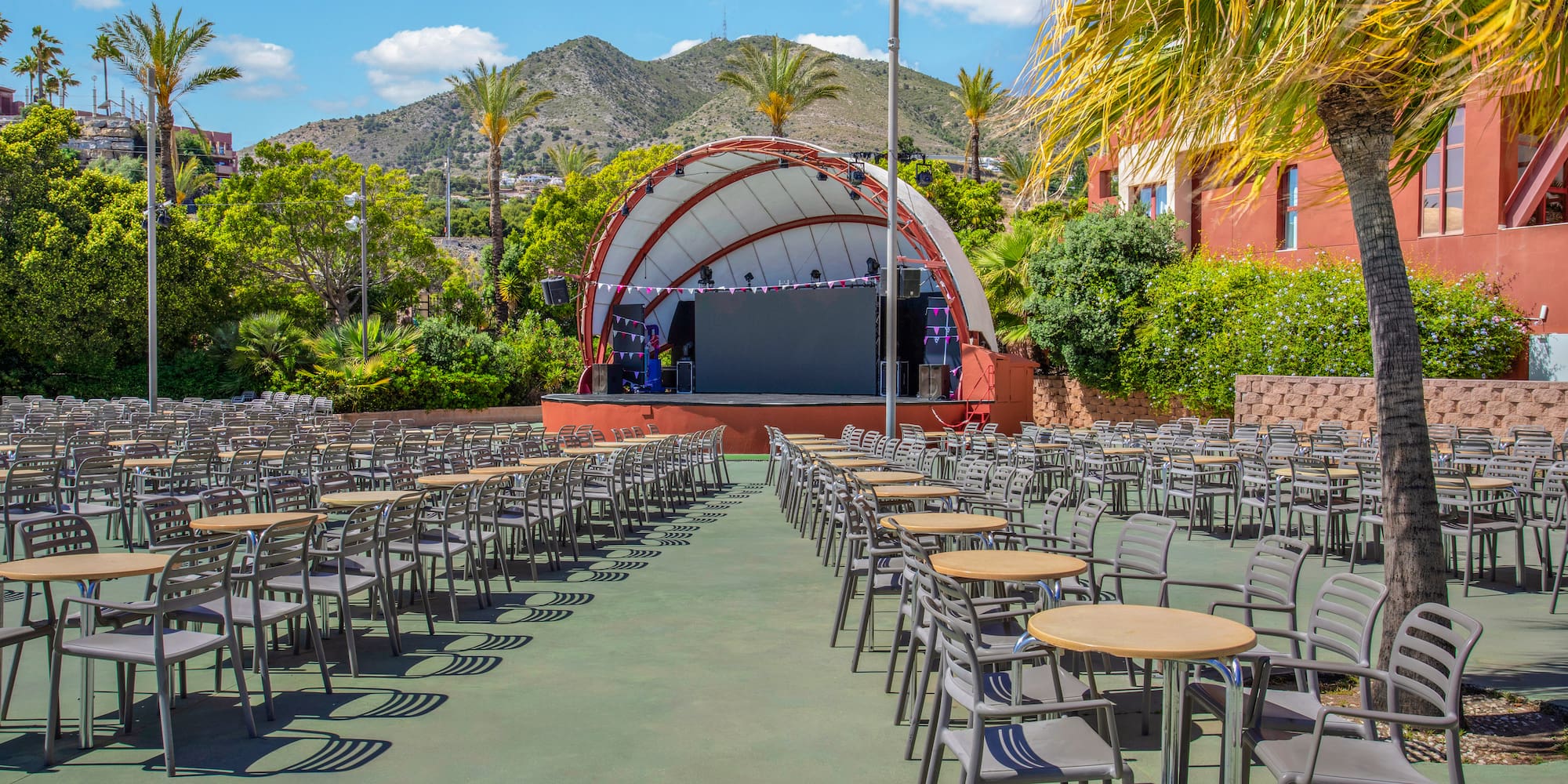 Sunny outdoor amphitheater with stage, rows of seating, tables, palm trees, mountains.