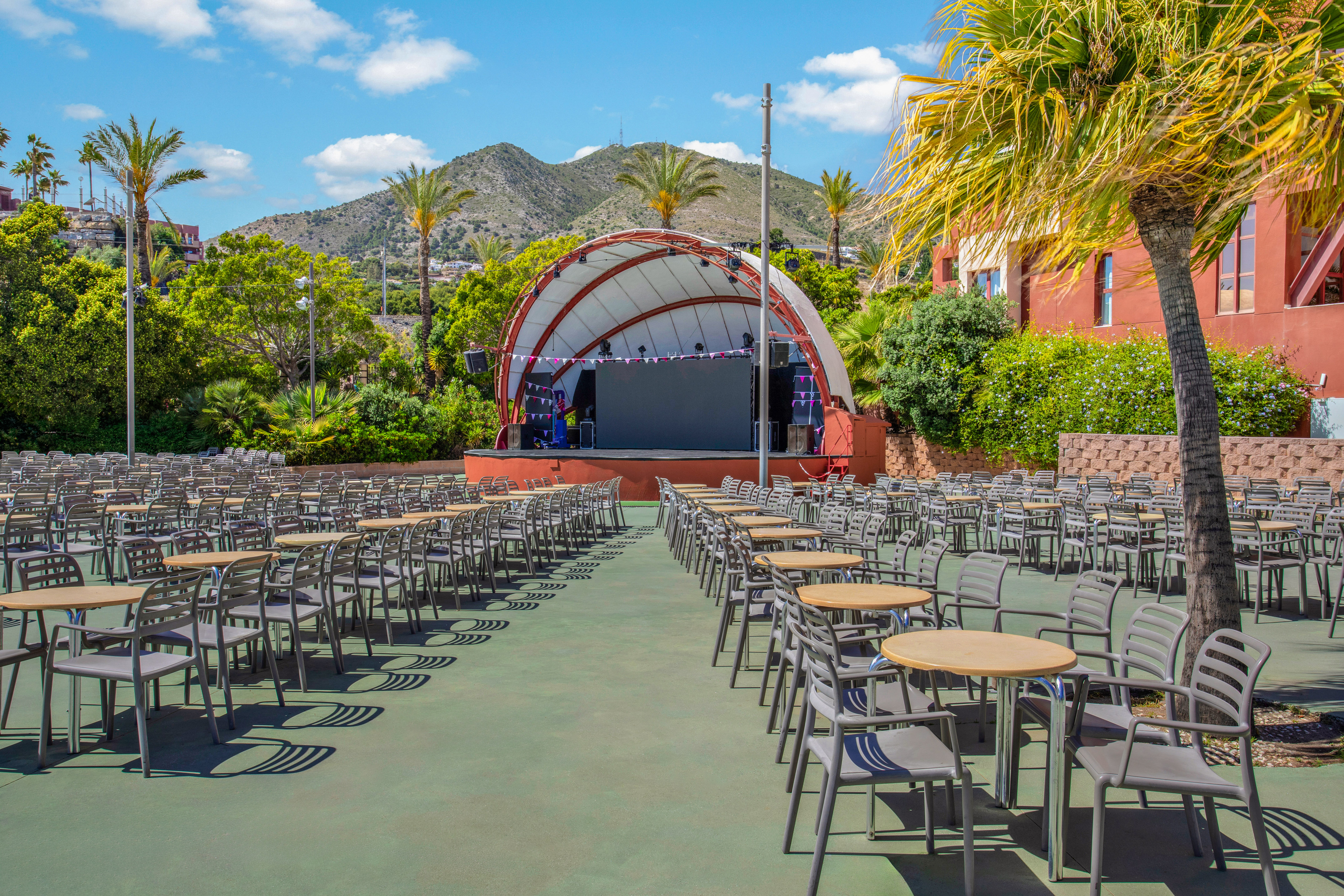 Sunny outdoor amphitheater with stage, rows of seating, tables, palm trees, mountains.